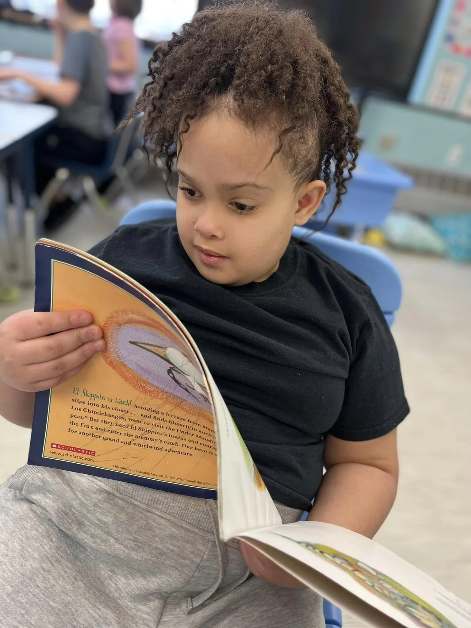 Young boy with curly hair reading a book in a classroom setting.