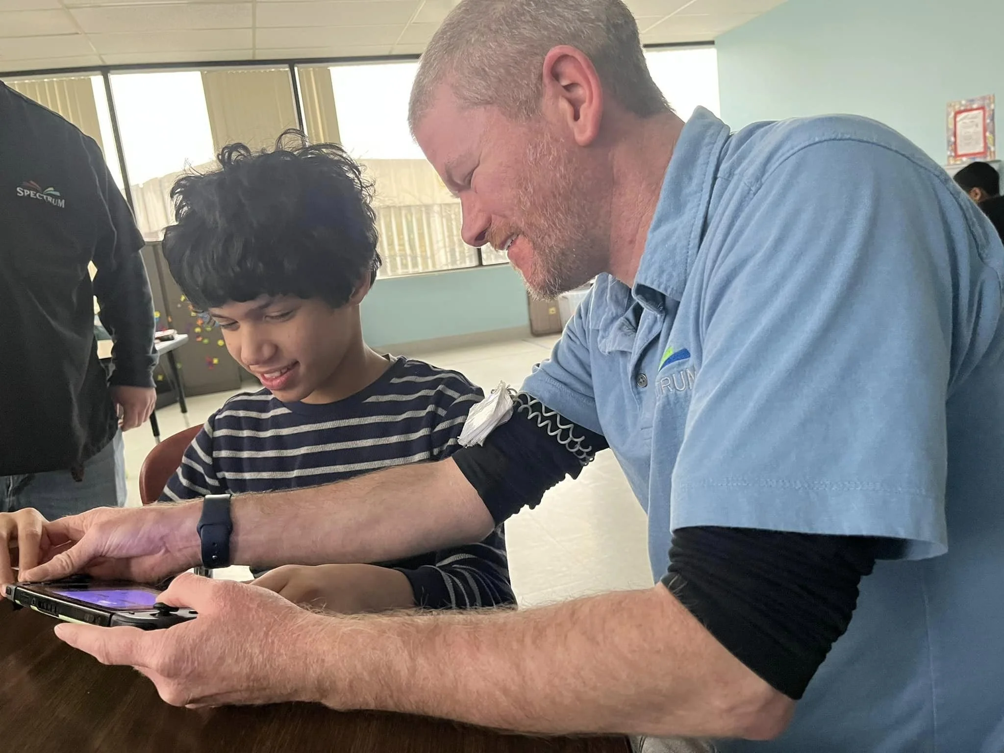 A man and a boy looking at a smartphone together in a room with large windows. The man is smiling and the boy has a concentrated expression.