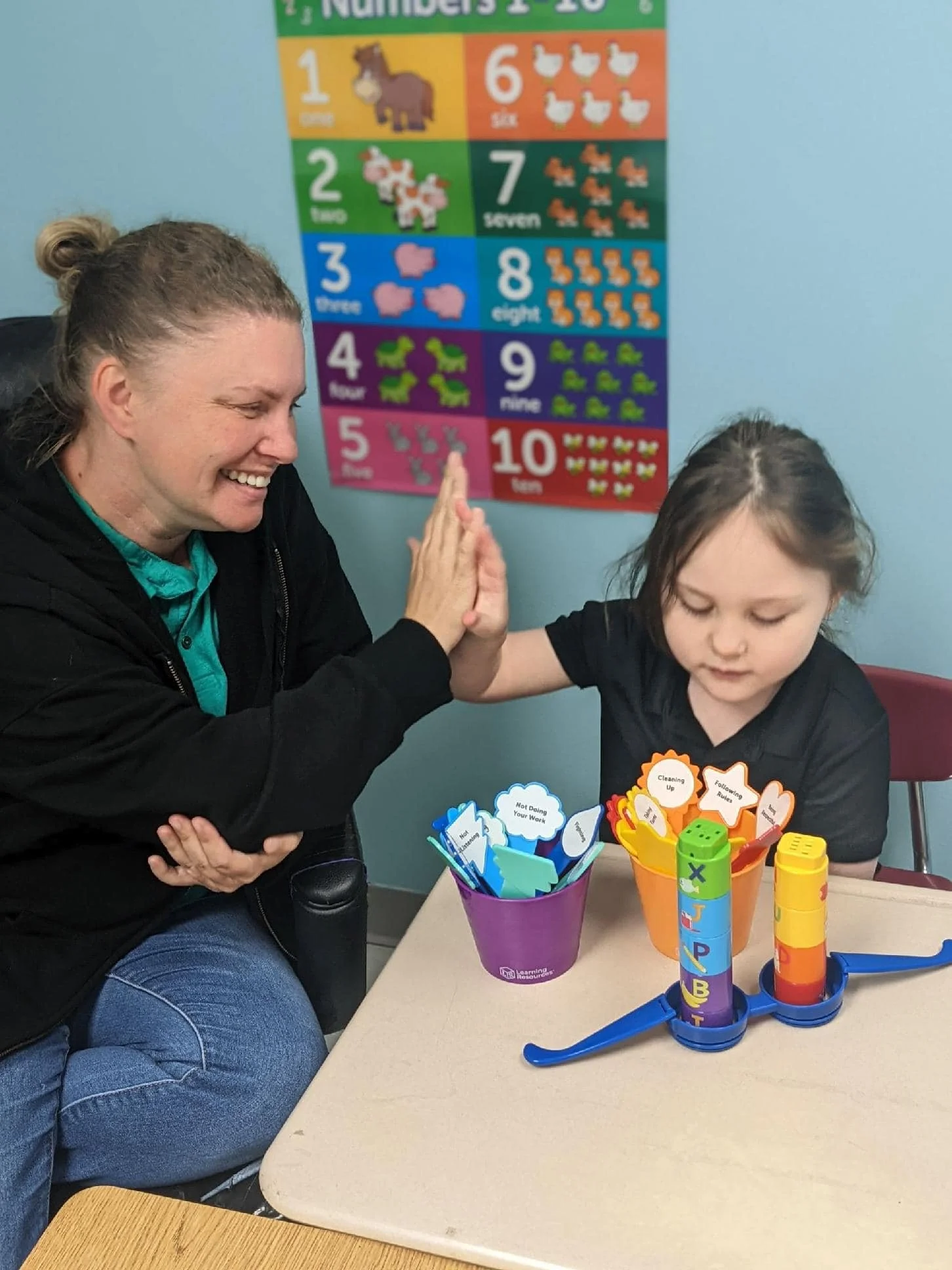 A woman and a young girl give each other a high five at a table in a classroom, with colorful educational posters and learning tools around them.