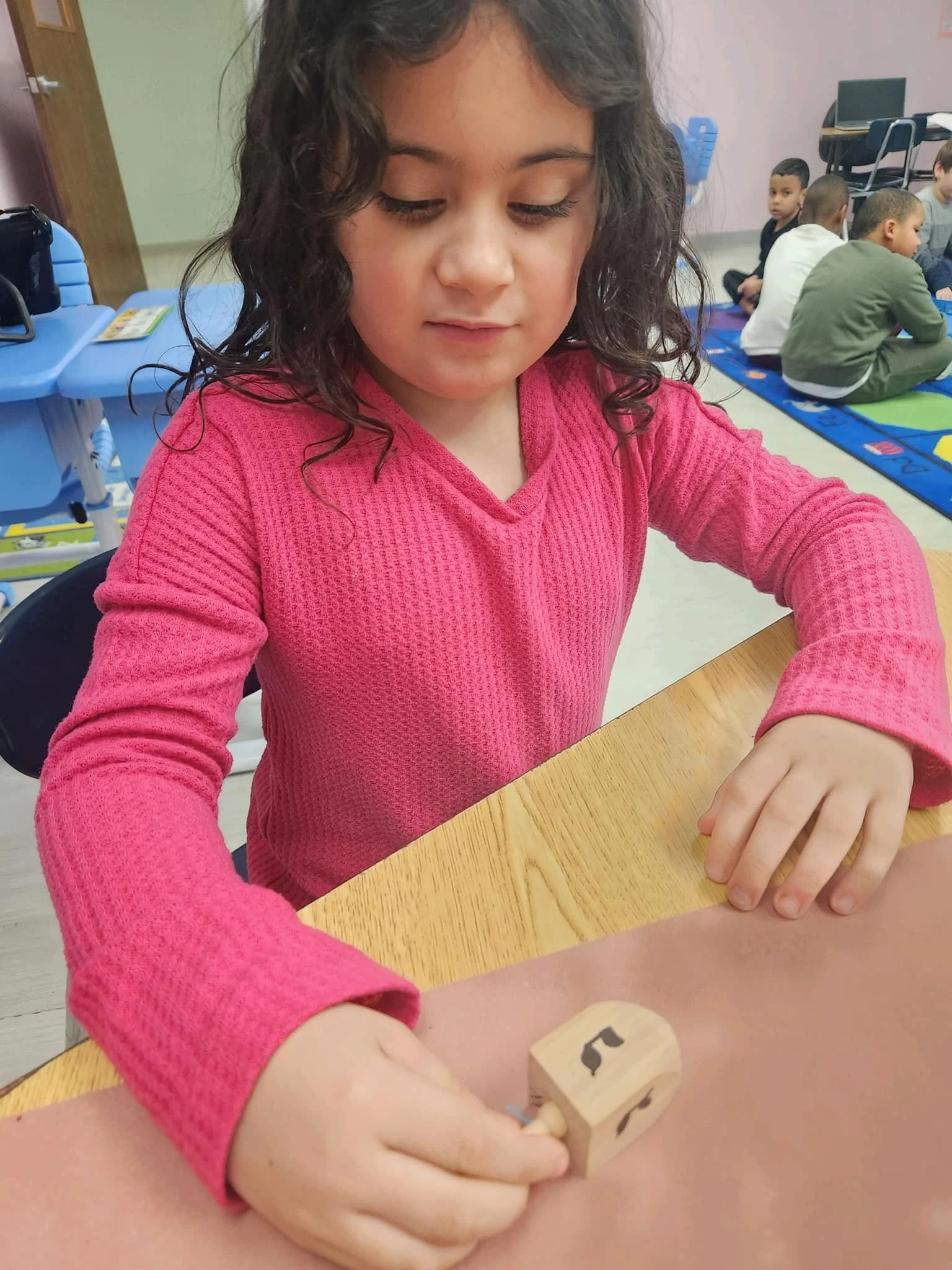 A young girl with curly dark hair wearing a pink sweater sitting at a wooden table, holding a wooden cube with black symbols. In the background, children are sitting on a colorful mat in a classroom setting.