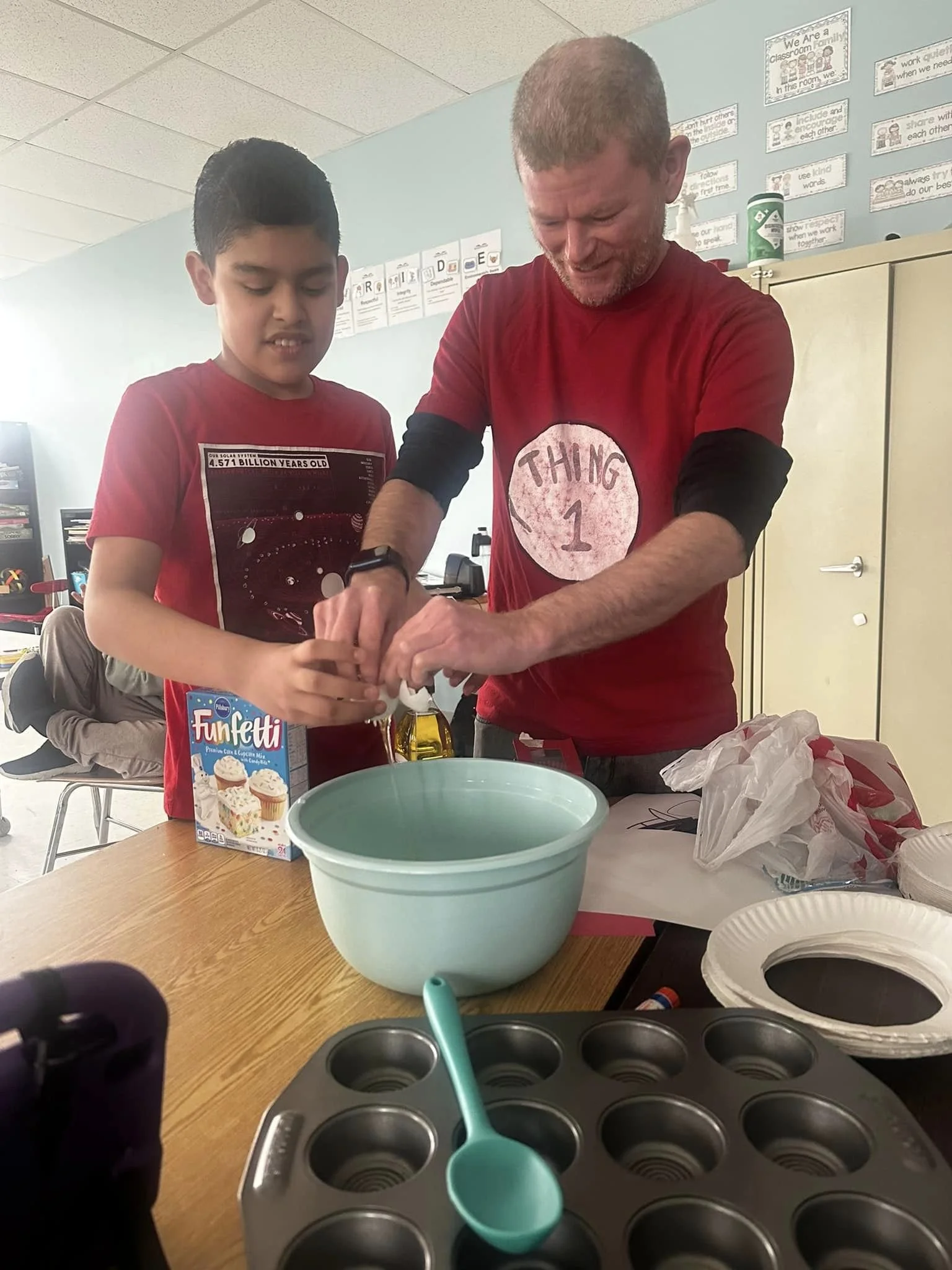 A man and a boy are baking together at a table in a classroom. They are cracking eggs into a bowl, with baking supplies around them, including cupcake liners and a muffin tin. The man wears a red shirt with 'Thing 1' on it, and the boy wears a red t-shirt with a space-themed design.