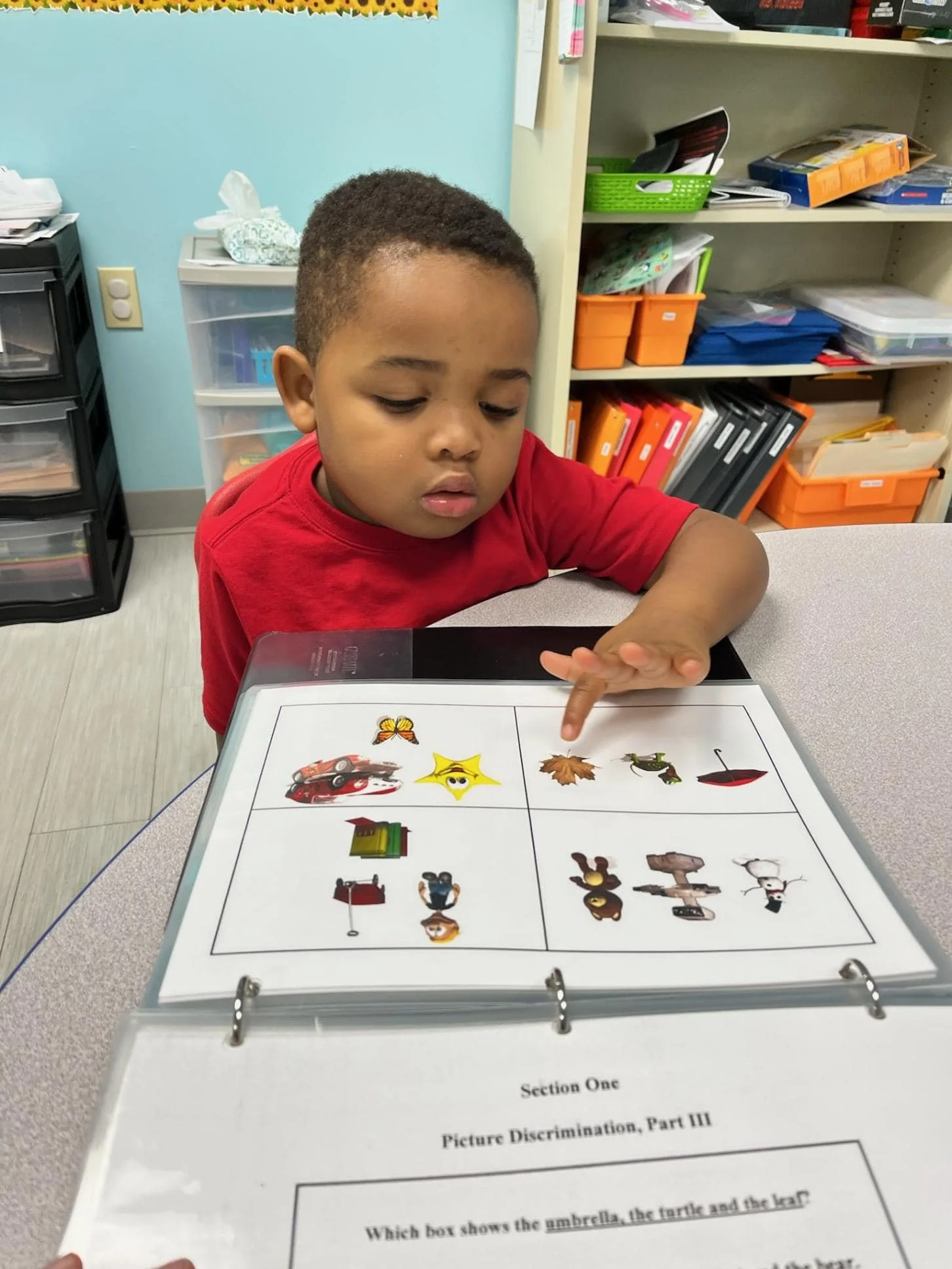 A young boy in a red shirt looking at a laminated sheet with pictures divided into four sections, holding up his finger, in a classroom with shelves and storage bins in the background.