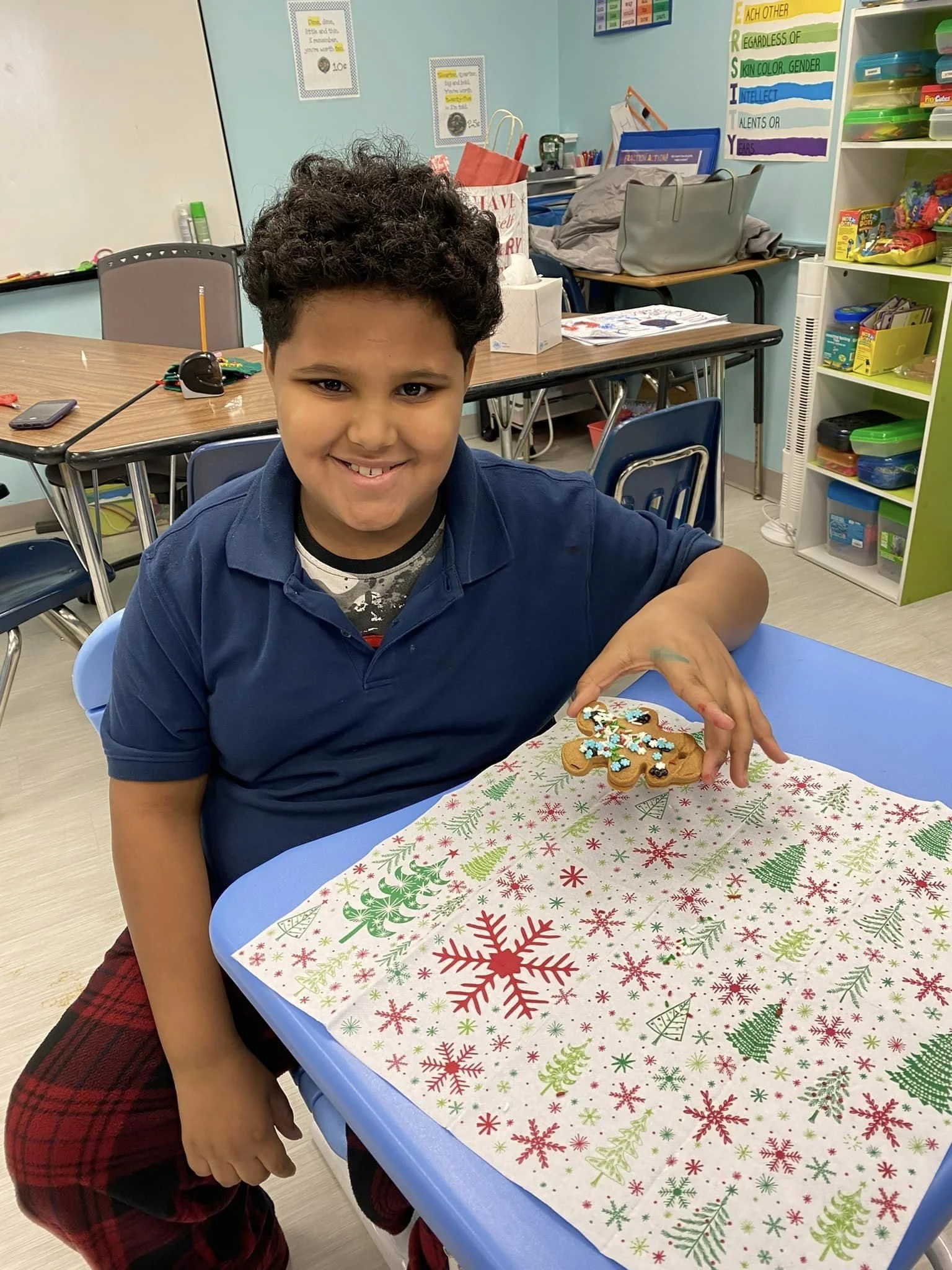A young boy smiling while sitting at a table with a decorated Christmas cookie in his hand. The tablecloth has a festive holiday pattern with green and red snowflakes and trees, and the background shows classroom supplies and colorful educational posters.