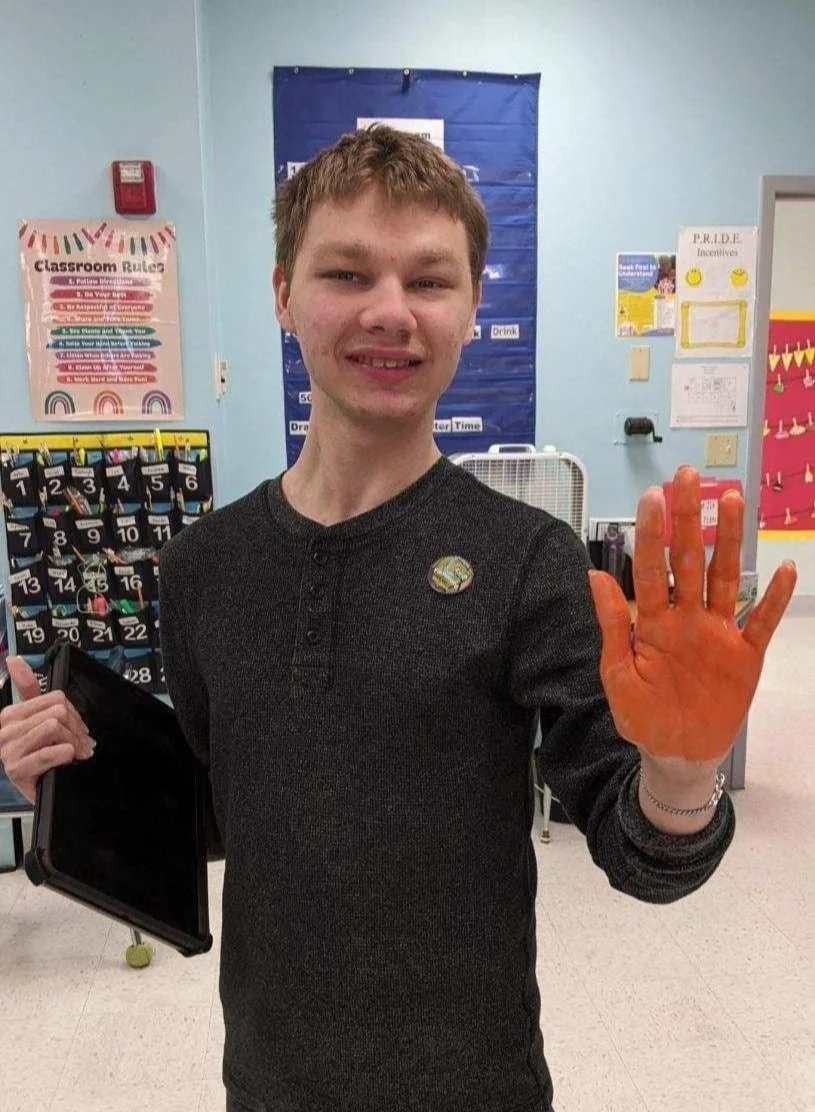 A young man in a classroom, smiling and raising his right hand with a painted orange palm. He is holding a tablet in his left hand and wears a round pin on his dark sweater. The classroom has educational posters and lockers in the background.