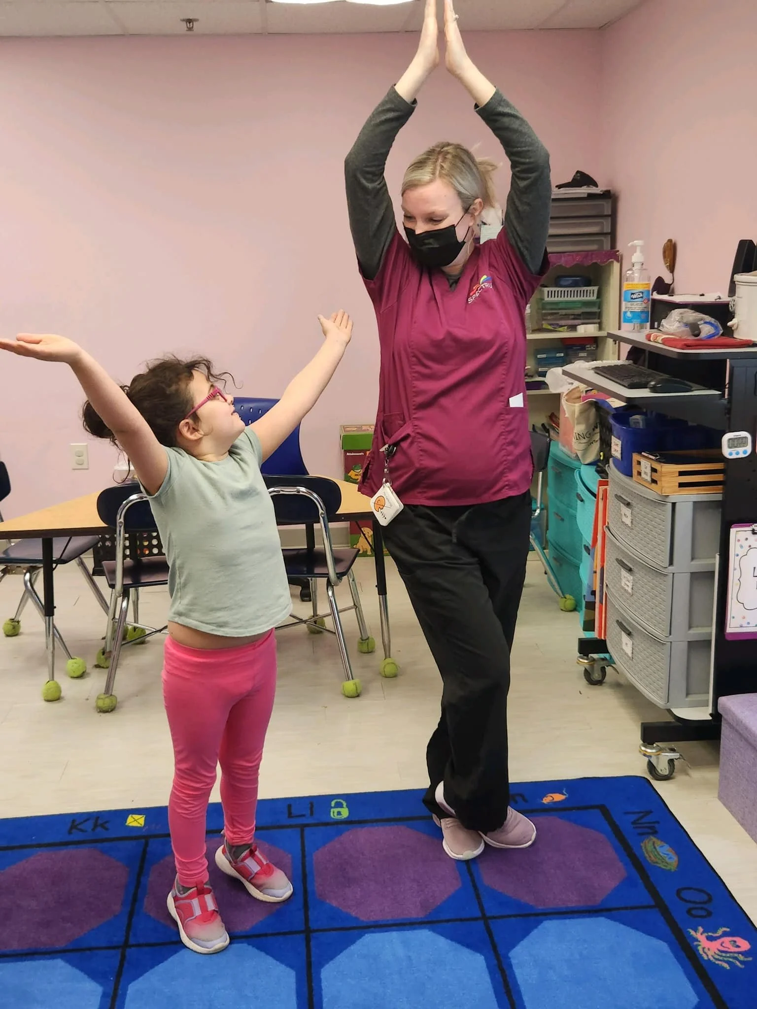 A young girl and a woman, likely a teacher or therapist, are standing on a colorful alphabet rug in a classroom. The girl has her arms stretched out and is reaching towards the woman, who has her hands raised above her head in a prayer-like pose. Both are smiling and appear to be engaged in a playful activity.