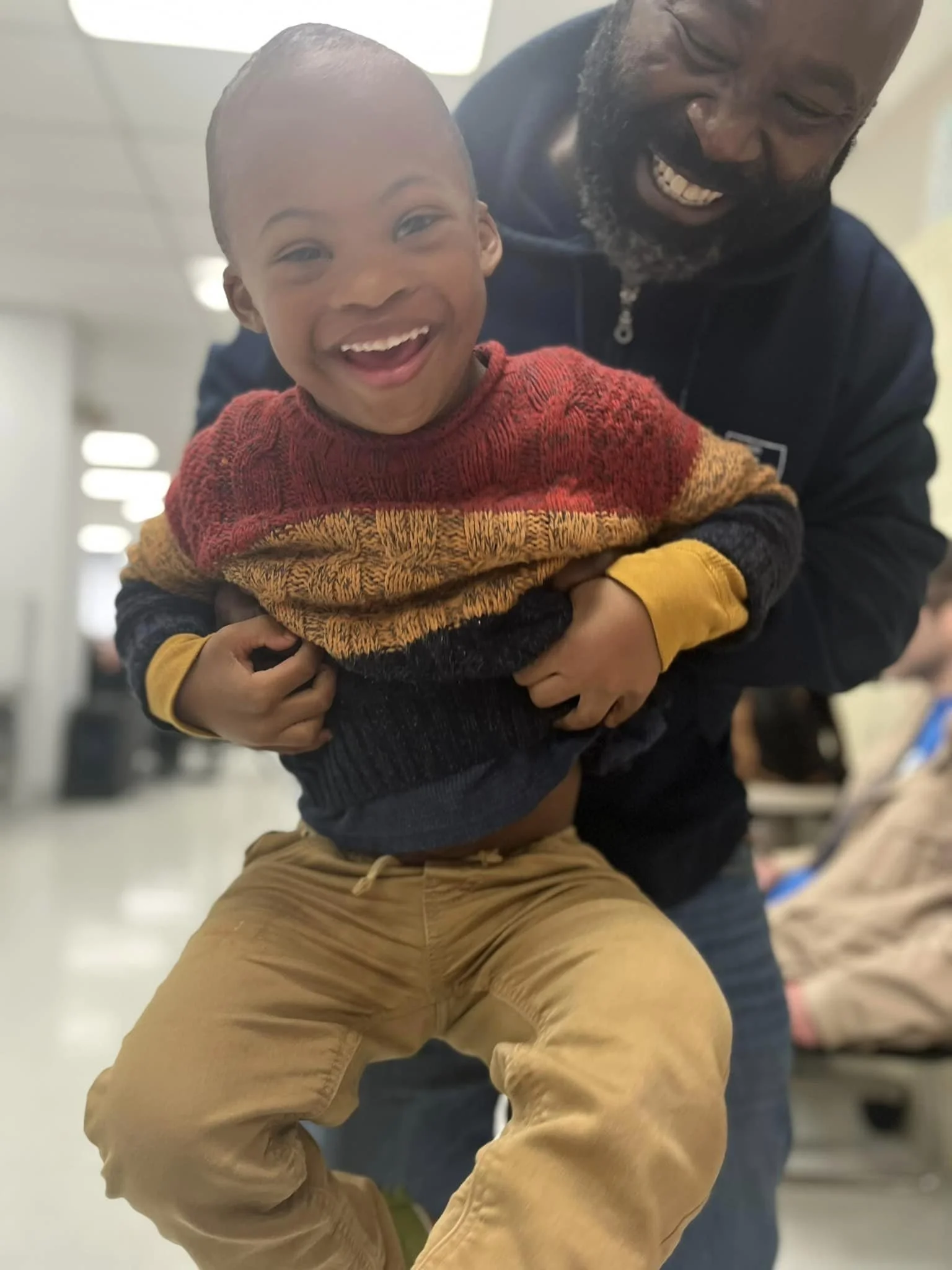 A young boy and an adult man smiling together in an indoor setting, with the man holding the boy up as they both enjoy playing or sharing a joyful moment.