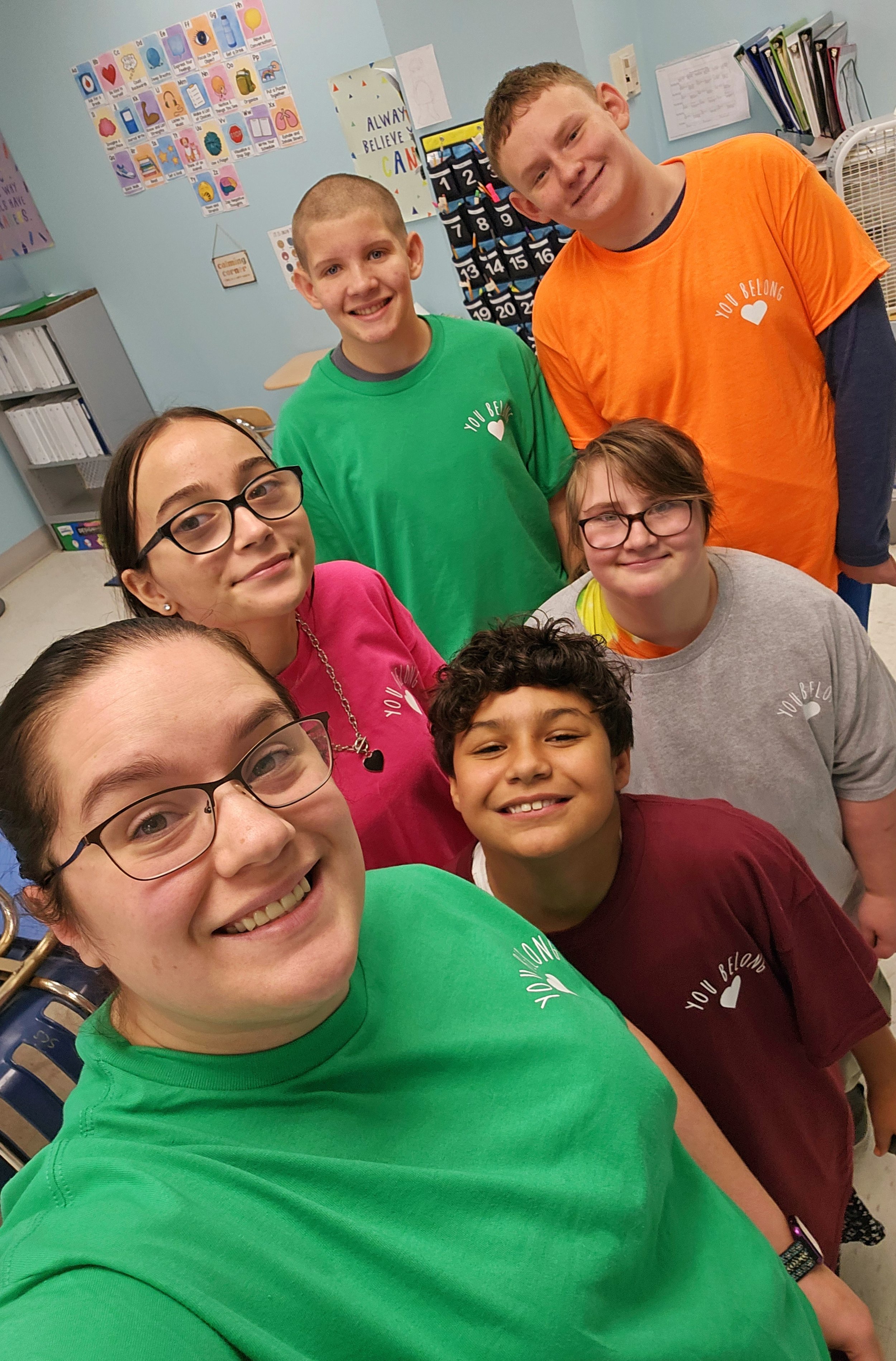 Group of seven children and a young woman taking a selfie in a classroom, all wearing colorful t-shirts with a heart and words 'You Belong' on them.