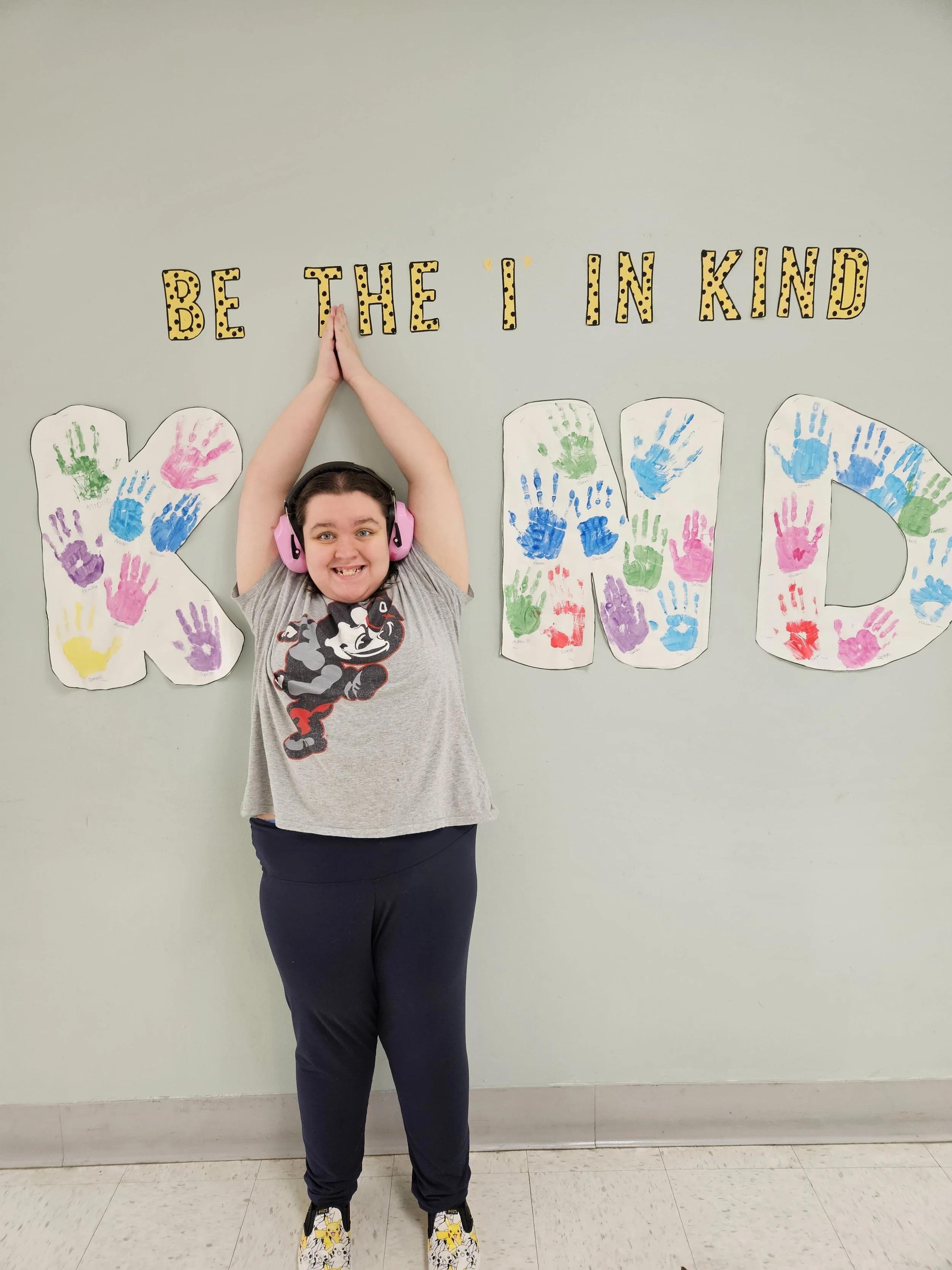 A young girl standing in front of a wall decorated with colorful handprints and large letters spelling 'KIND'. The wall also has a banner that says 'BE THE I IN KIND', and the girl is smiling while wearing pink headphones, a gray Minnie Mouse t-shirt, black pants, and sneakers with a Minnie Mouse design.
