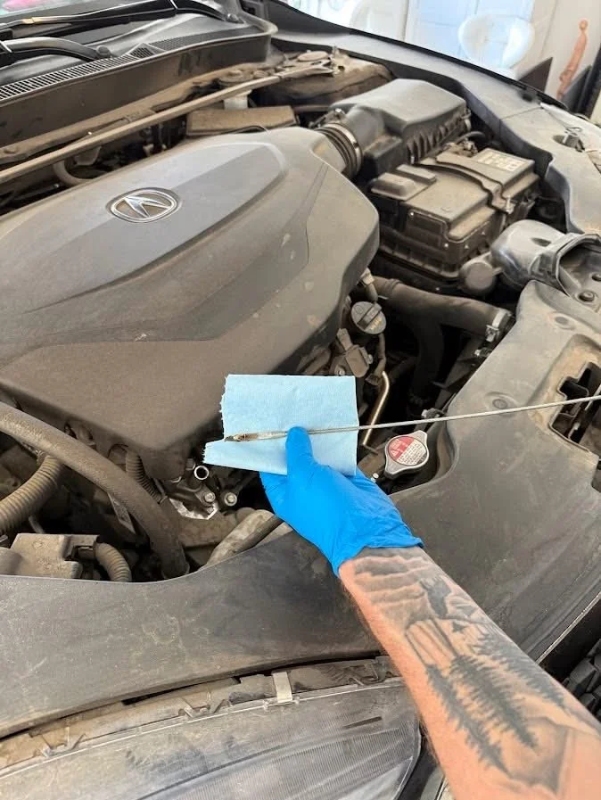 A person wearing a blue glove and a tattooed arm is holding a dipstick with a blue paper towel wrapped around it, checking the oil level in a car engine.