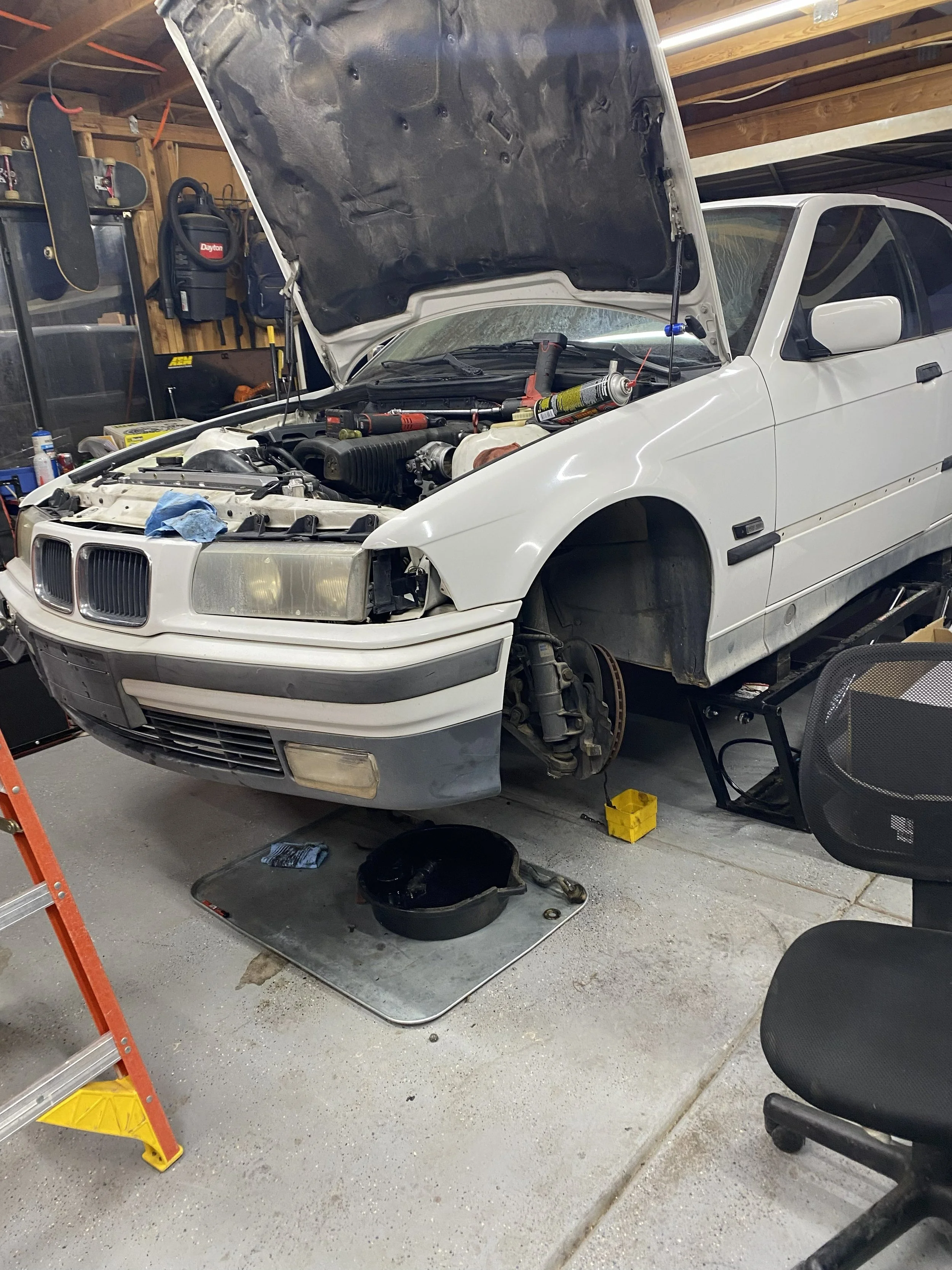 A white car is being repaired in a garage with its hood open. The front bumper is removed, exposing the engine. Various tools and parts are scattered around, including a black pan, a yellow tool box, and a black chair.