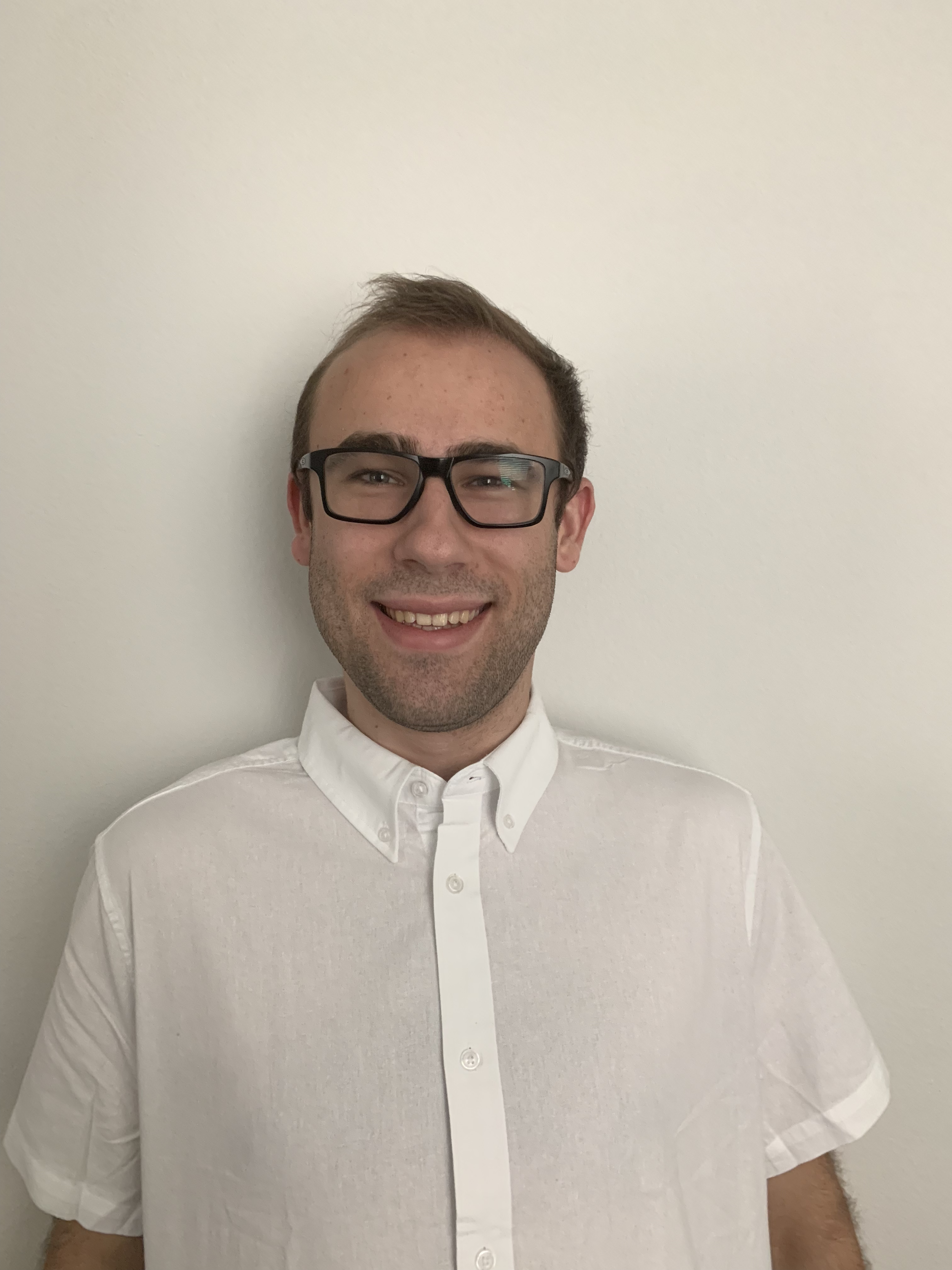 A young man with short brown hair, wearing glasses and a white button-up shirt, smiling and standing against a plain white wall.