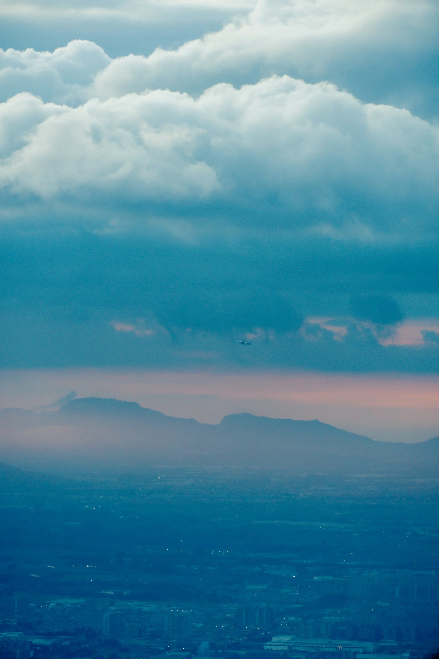View of a cityscape from above with mountains and clouds in the background, and a small airplane flying in the sky.
