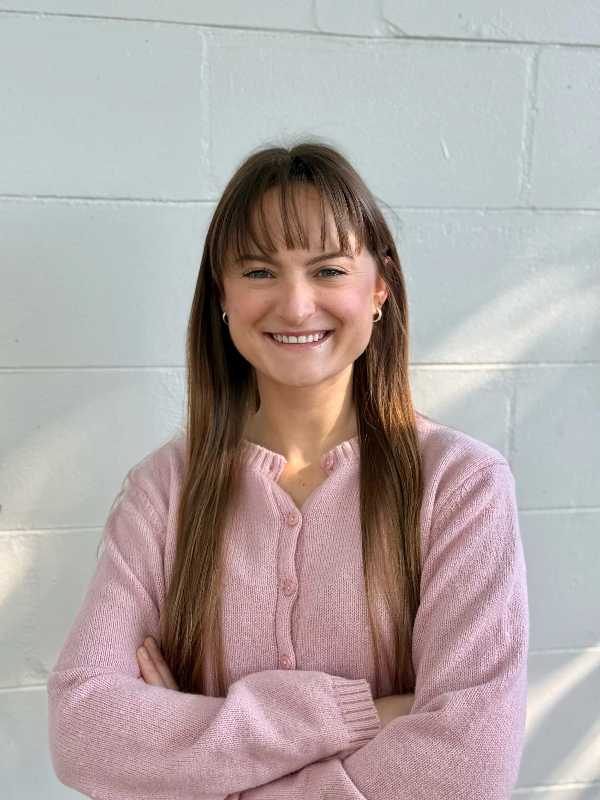 A woman with long brown hair and bangs smiling with arms crossed, wearing a pink button-up cardigan, standing against a light-colored wall.
