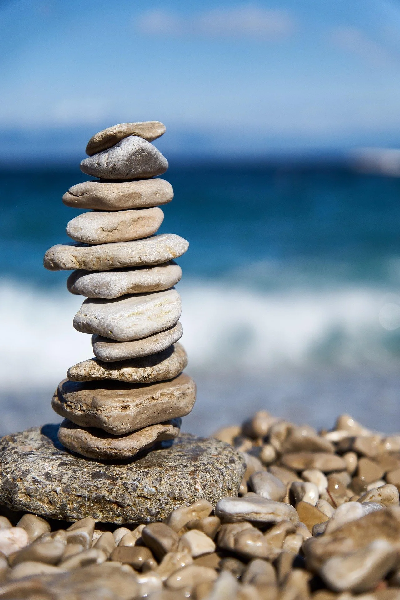 Stack of flat stones balanced vertically on a larger rock at a beach, with ocean and sky in the background.