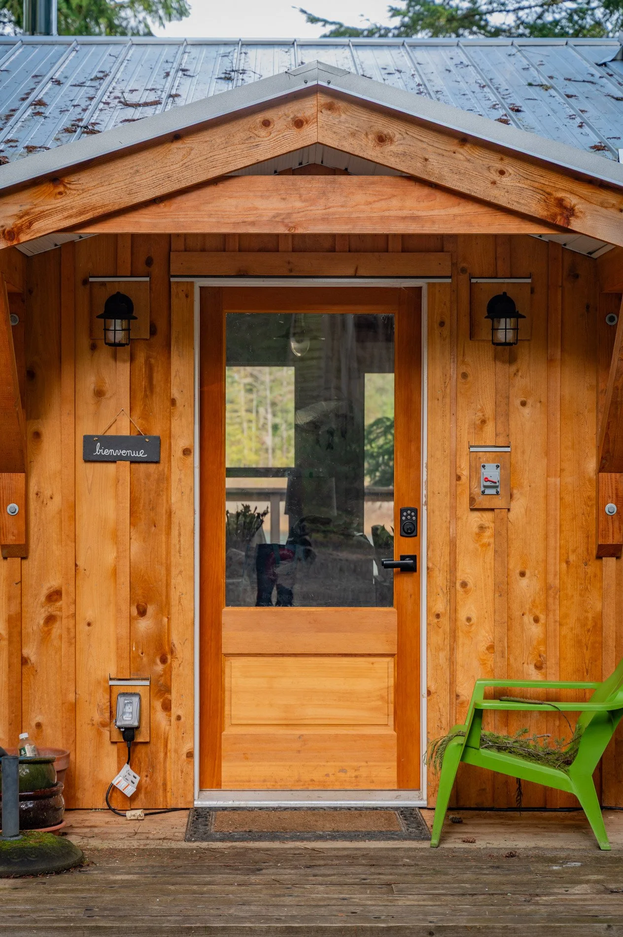 Front entrance of a wooden cabin with a glass-paneled door, a green chair with dried grass on it, and a welcome sign that says 'bienvenue'.