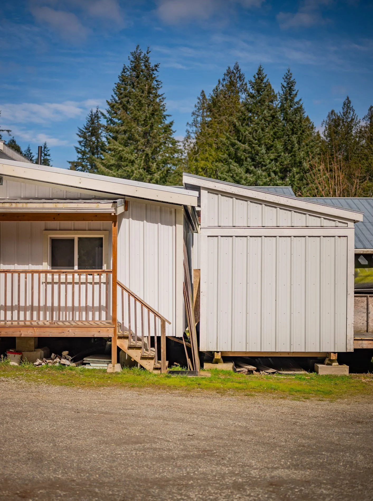 White house with wooden porch and stairs, exterior showing siding, surrounded by tall pine trees under blue sky.