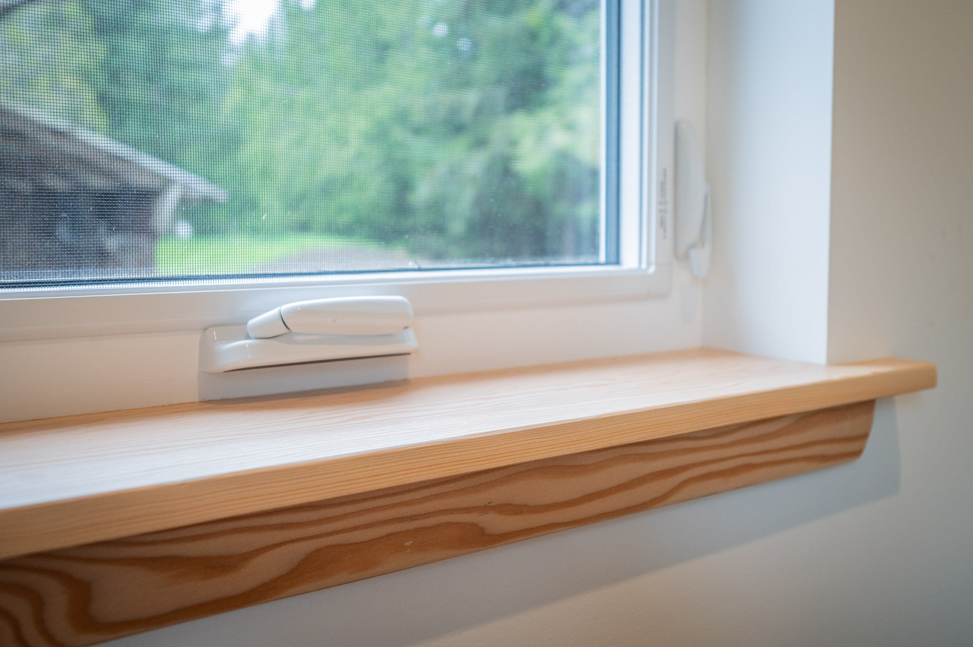 A window with a white frame and a tilt latch, looking out onto a green outdoor area, with a wooden window sill beneath.