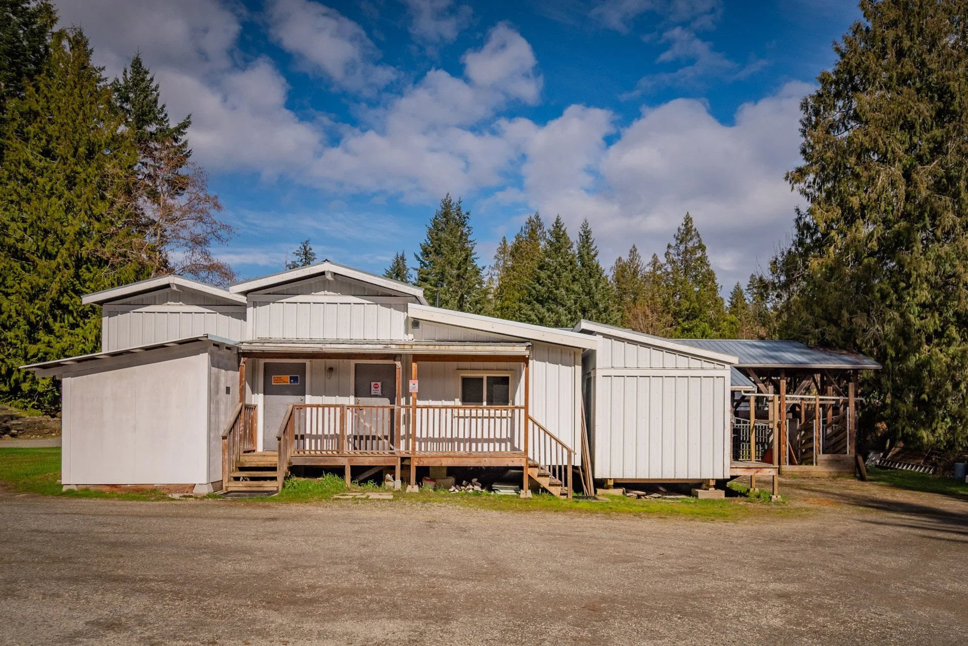 A white metal building with a small wooden porch, surrounded by tall green trees and a partly cloudy blue sky.