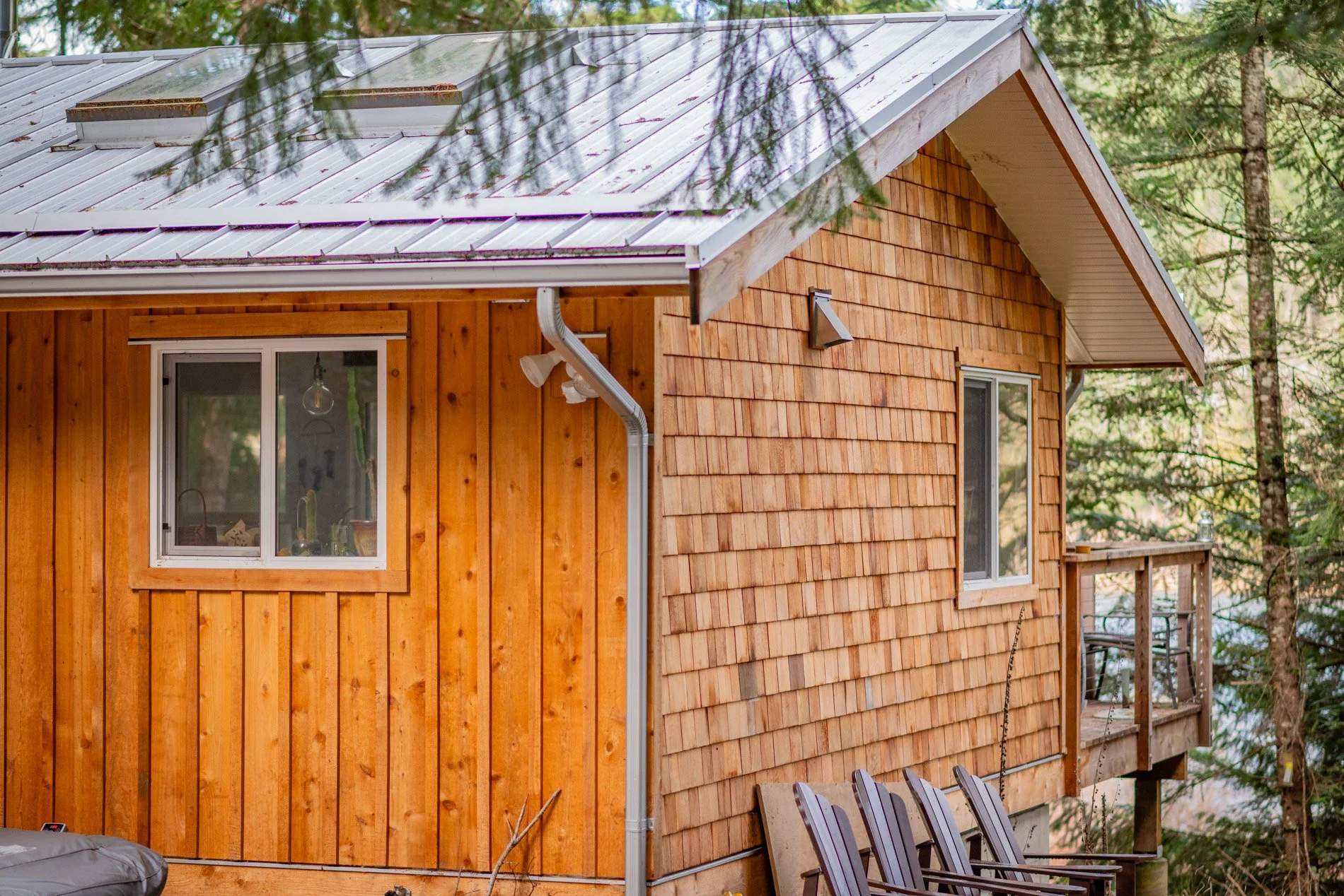 Side view of a wooden house with a metal roof, surrounded by trees, featuring a small balcony with a table and chairs.