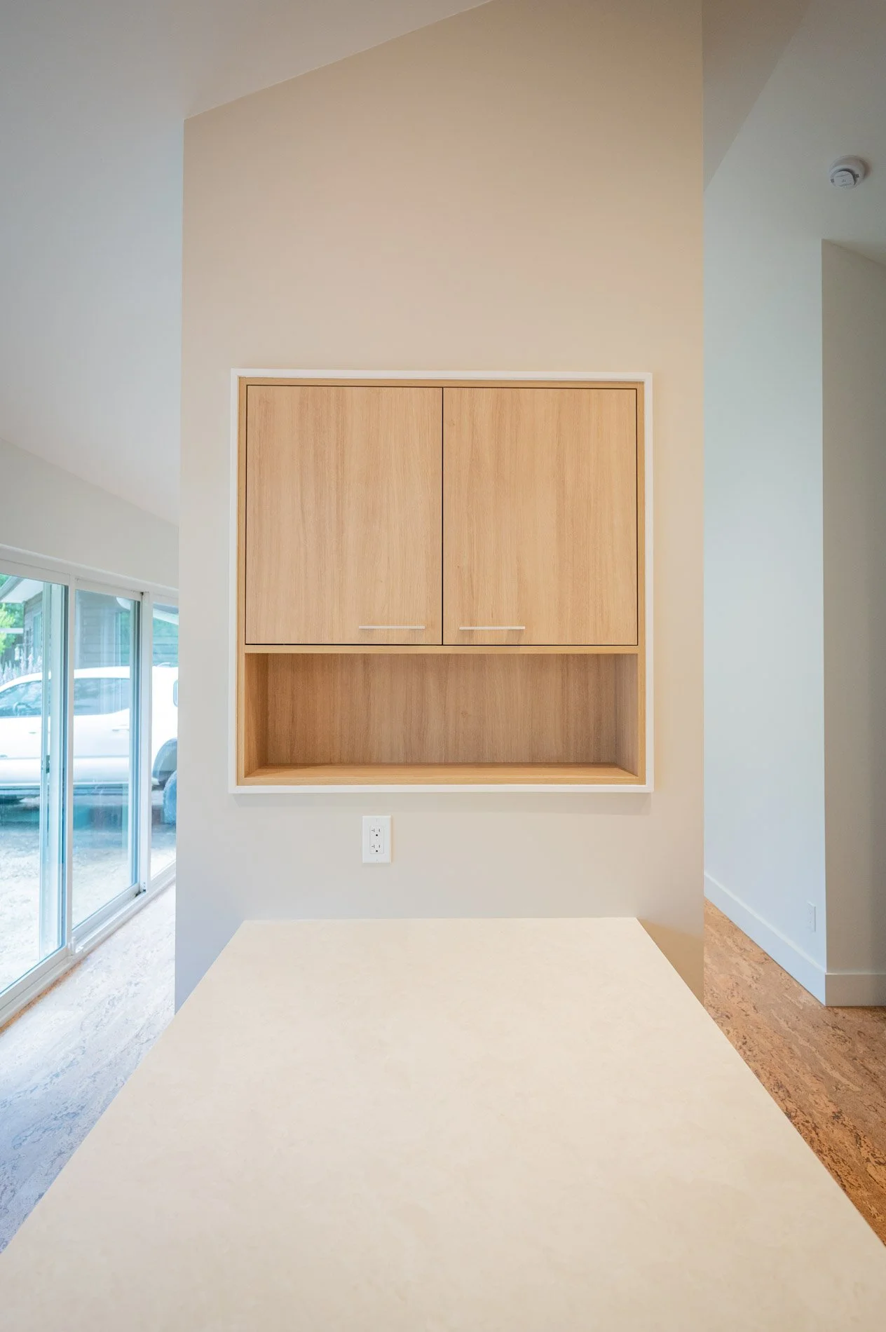 Empty built-in wooden cabinet with closed upper doors and an open shelf below, mounted on a beige wall in a modern room with large sliding glass doors and a view of a parked vehicle outside.