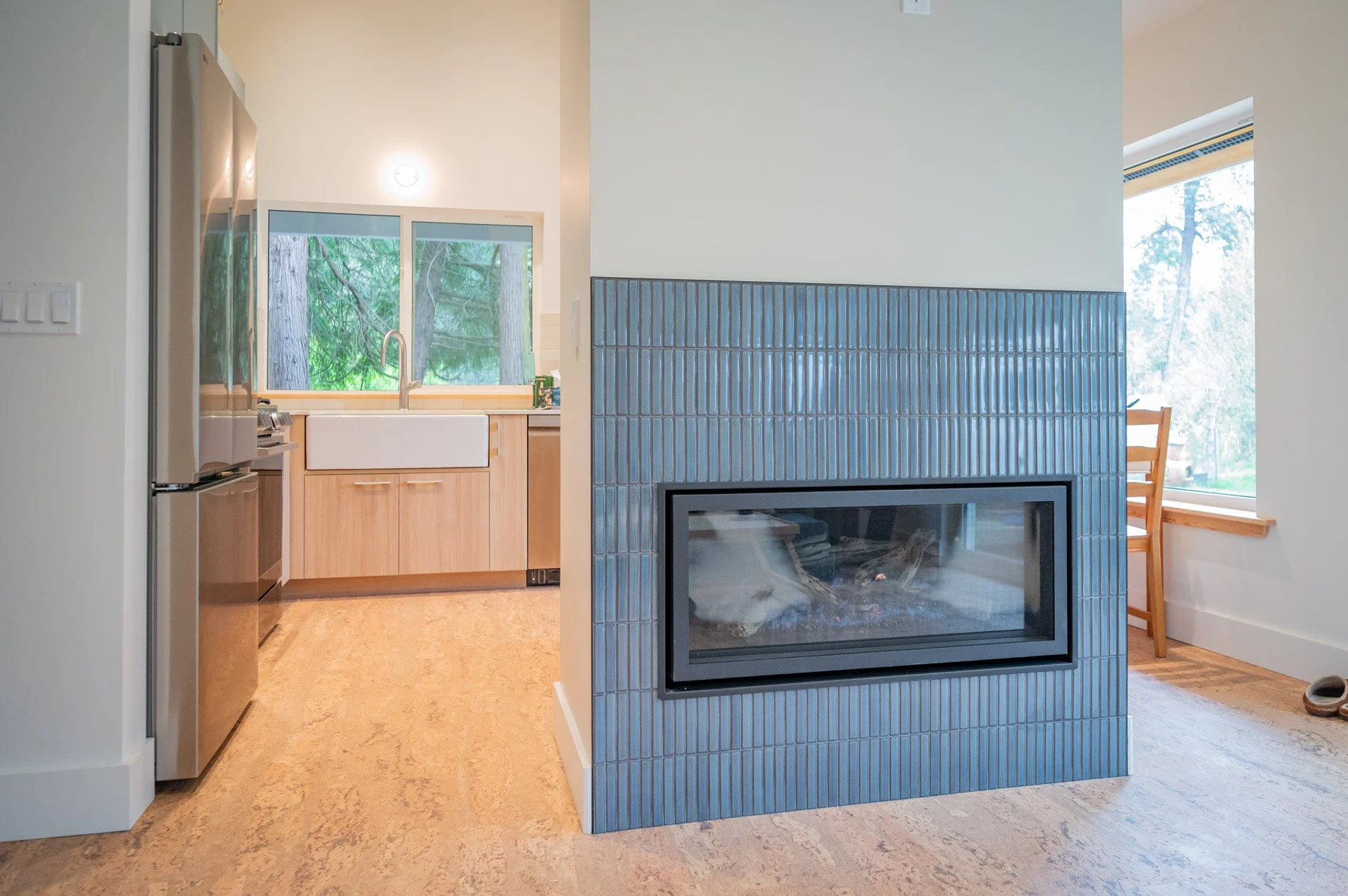 Interior view of a kitchen and a living area separated by a partial wall with a modern fireplace. The kitchen has wooden cabinets, a white farmhouse sink, and large windows showing trees outside. The living area has a window and a wooden chair near the window.