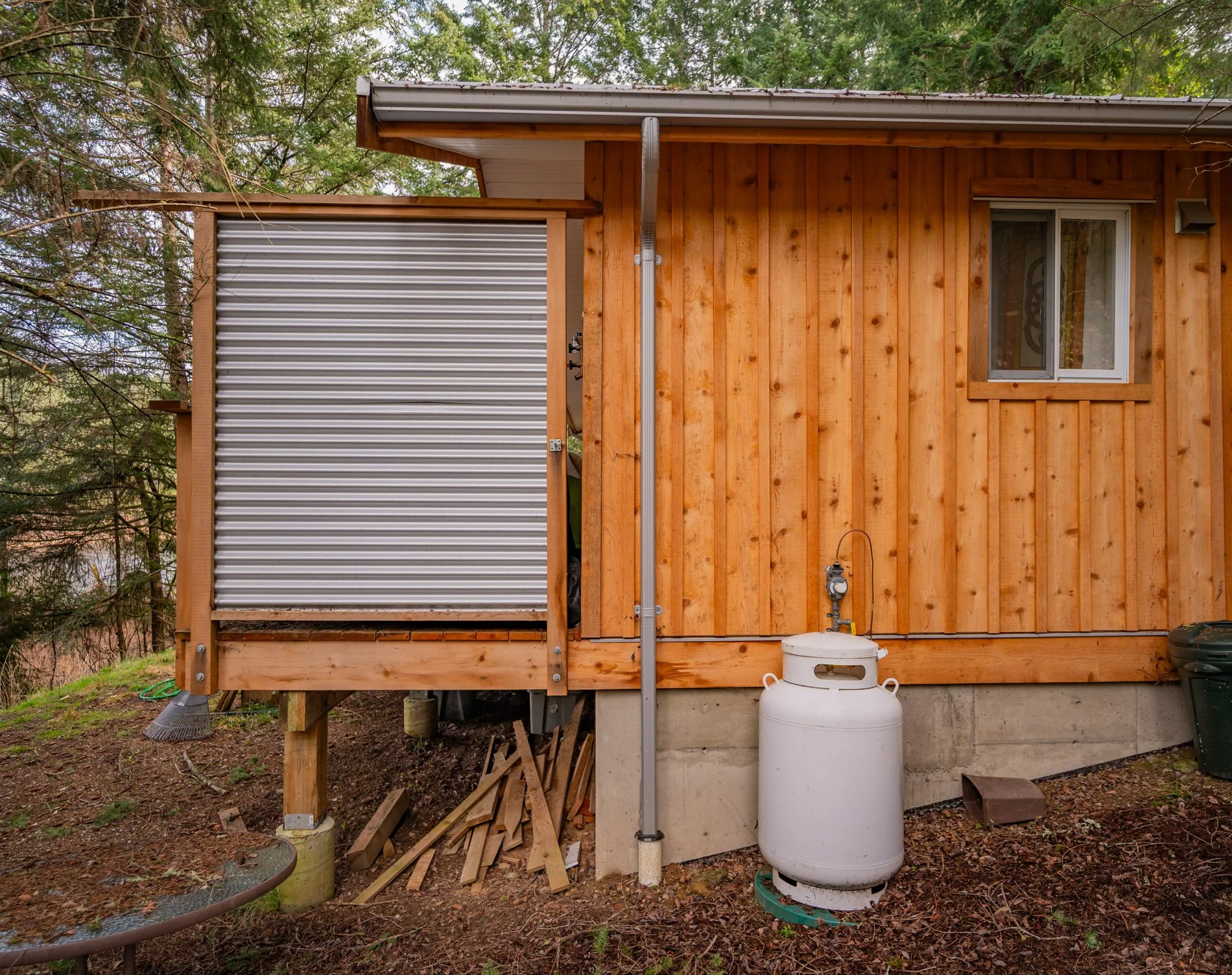 Back exterior of a wooden house with a small window, a propane tank, and a metal roll-up door.