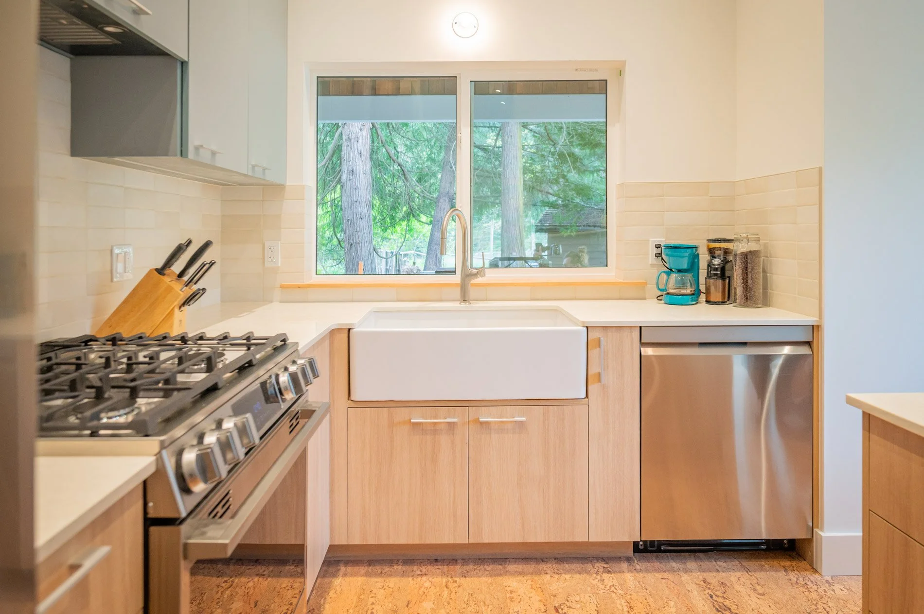 Modern kitchen with white sink, wooden cabinets, stainless steel dishwasher, gas stove, and window overlooking trees.