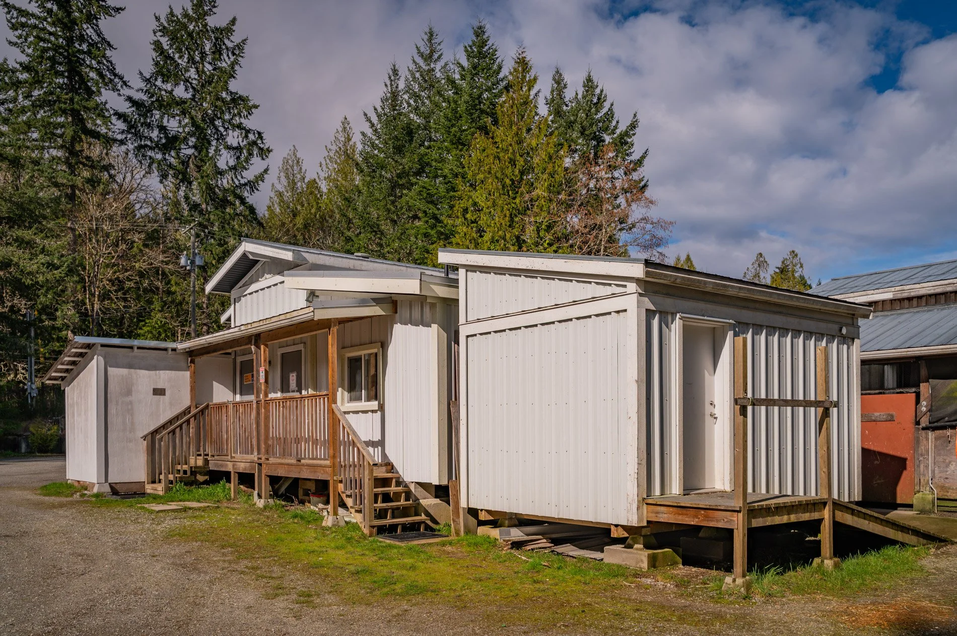 Buildings with white and gray siding, wooden steps and porch, surrounded by green grass and trees, under a partly cloudy sky.