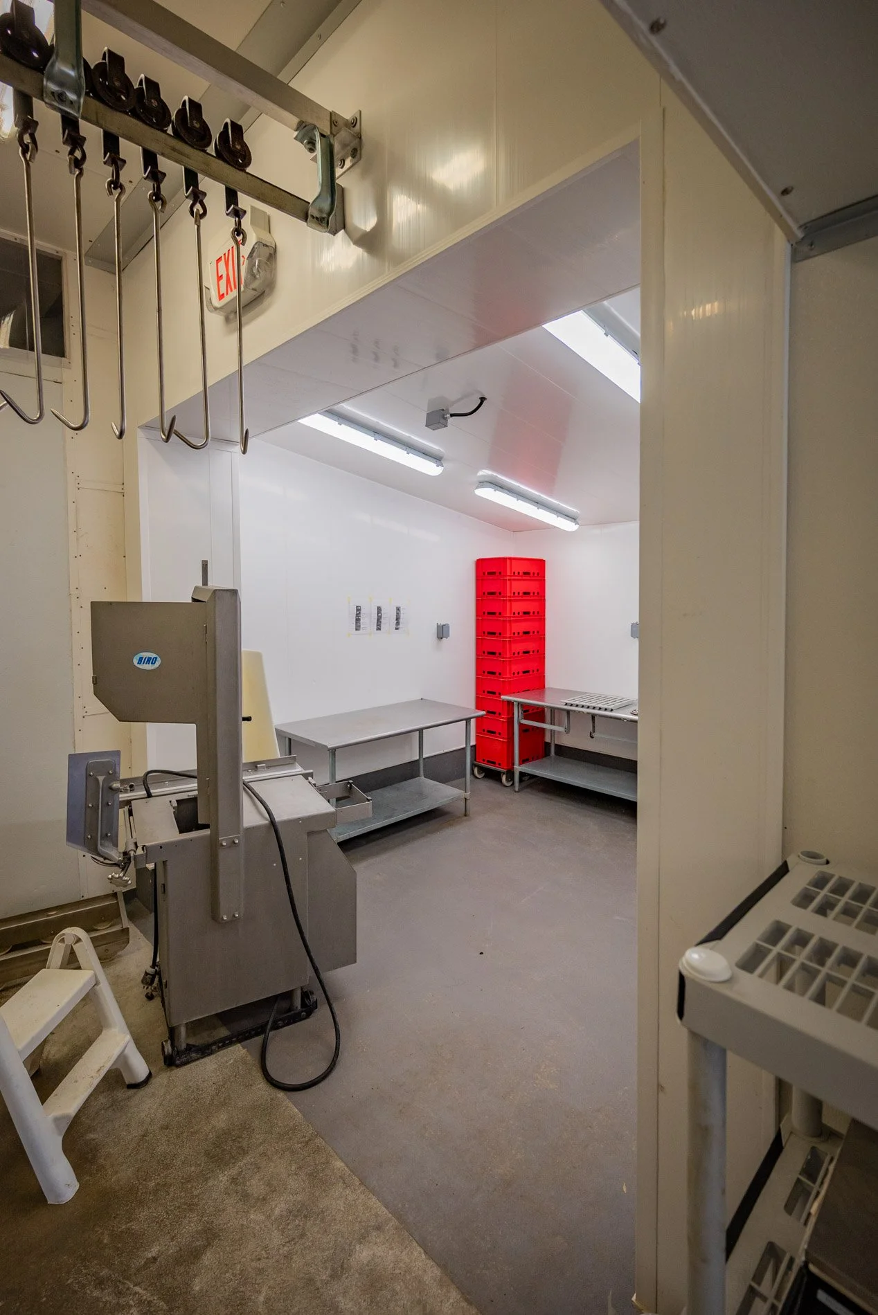 A small, sterile room with white walls and ceiling, illuminated by fluorescent lights, containing metal tables, red crates, and kitchen equipment.