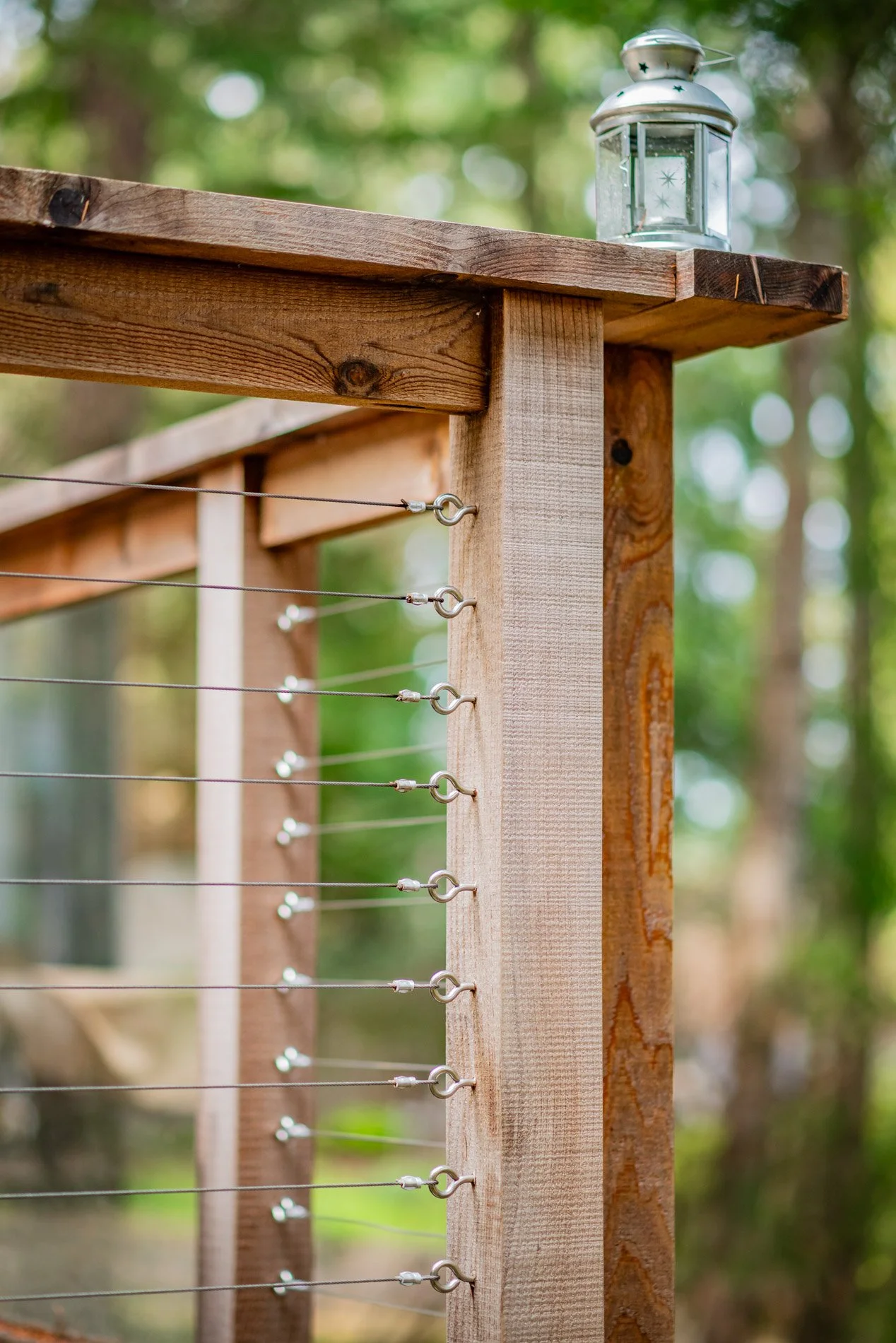Close-up of a wooden structure with metal wires attached to hooks, and a lantern placed on top, against a blurred green forest background.