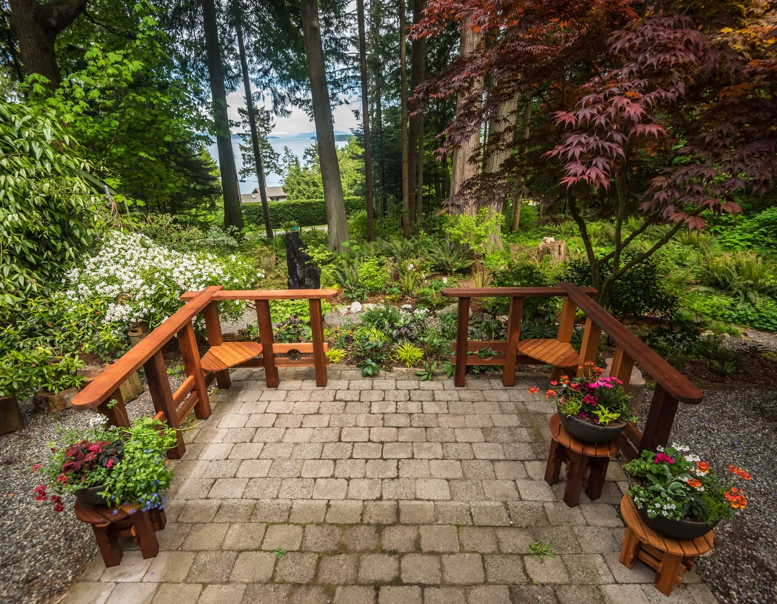 A cozy backyard patio with stone pavers, surrounded by lush greenery and flowering plants. There are wooden benches with potted flowers on top, and tall trees in the background with a view of water and islands in the distance.