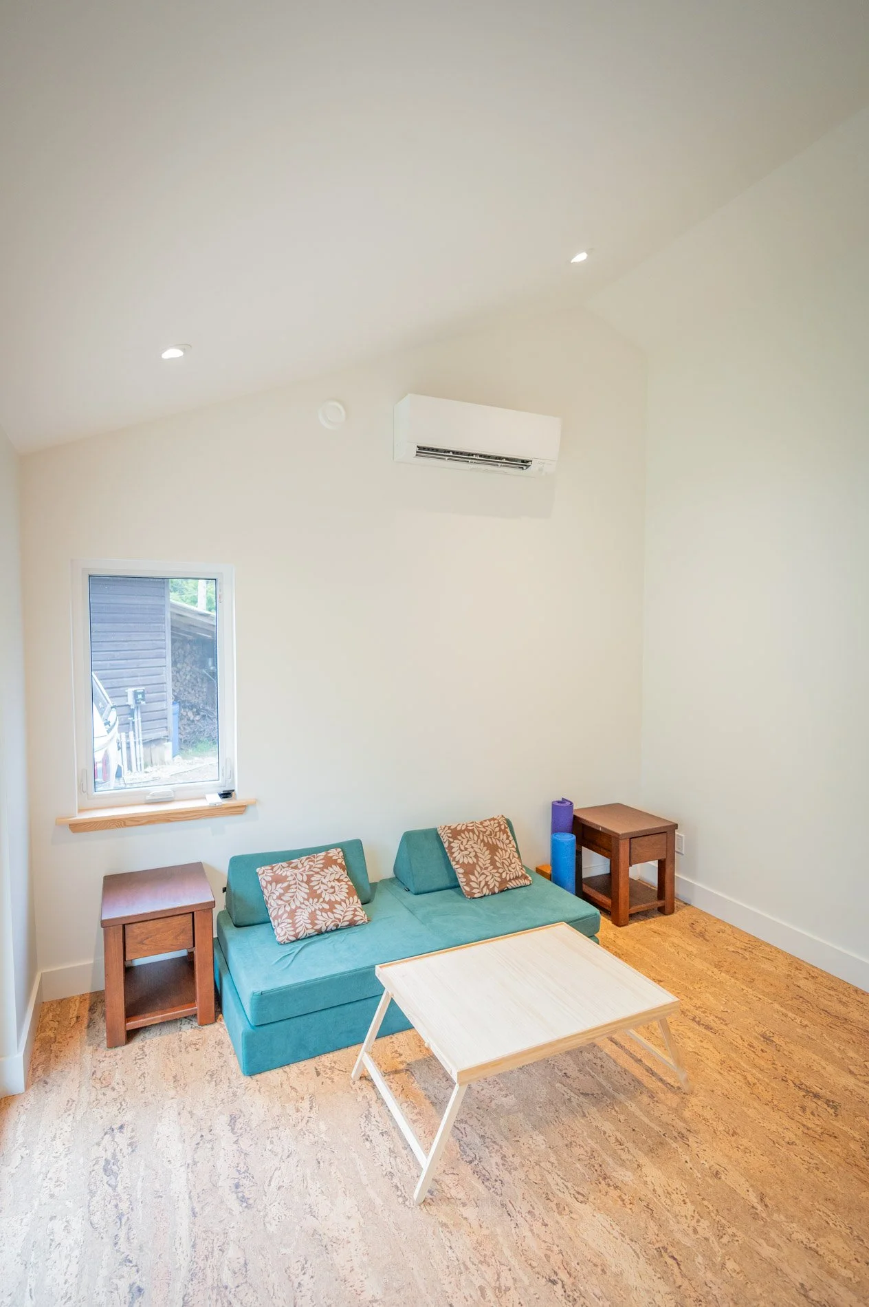 Living room with a turquoise sofa, two wooden side tables, a small wooden coffee table, a window, and an air conditioning unit on the wall.
