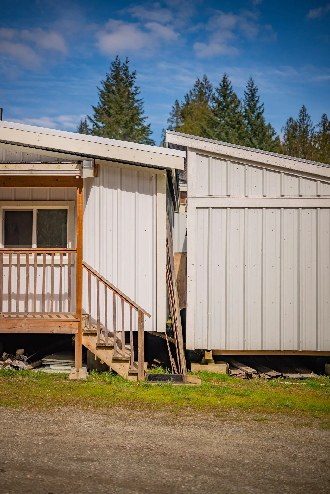 Two small white buildings made of metal siding, one with a wooden staircase and railing, situated on grassy and dirt ground, with trees and a partly cloudy blue sky in the background.