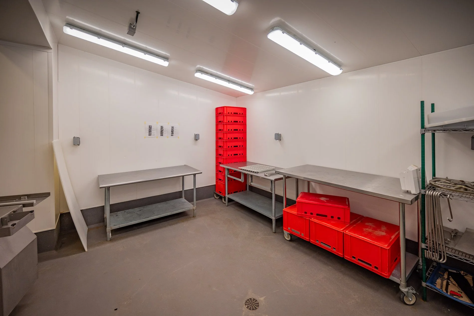 Empty industrial or commercial storage room with stainless steel tables, red plastic crates, a tall red storage rack, and metal shelving units.