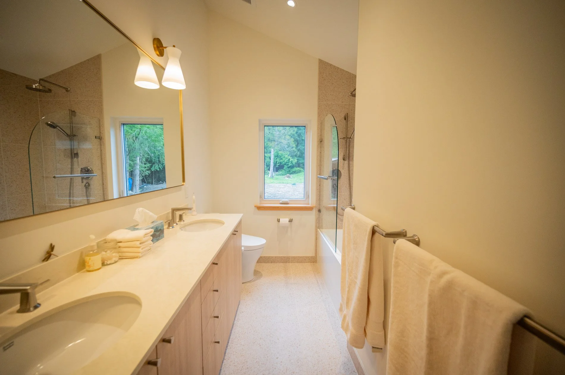 A bathroom with a double vanity sink, large mirror, shower with glass door, bathtub, and window showing outside greenery. Towel bars with towels and bathroom items on the counter.
