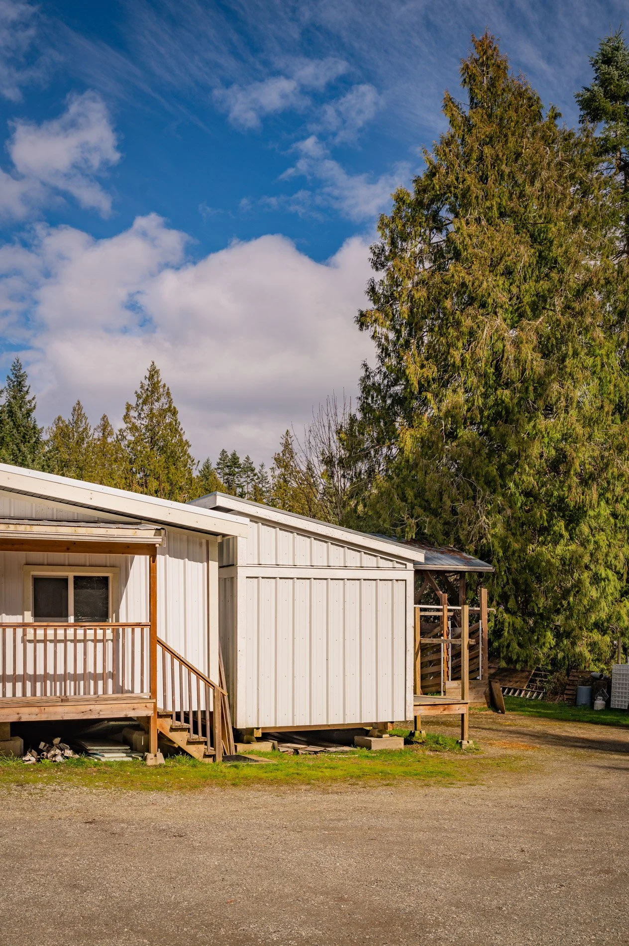 A small white building with a porch, surrounded by tall green pine trees under a partly cloudy blue sky.