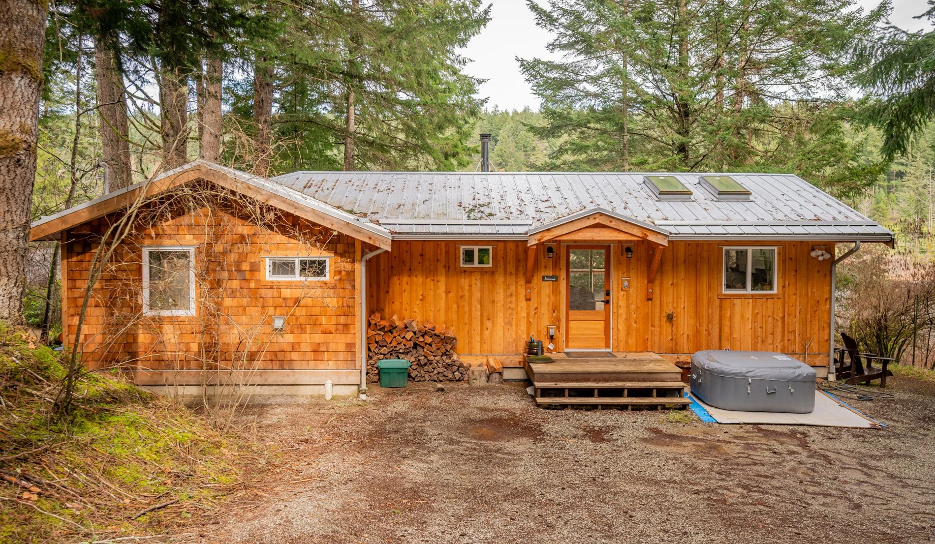 A small wooden house surrounded by trees with a metal roof, steps leading to the front door, and firewood stacked beside it.