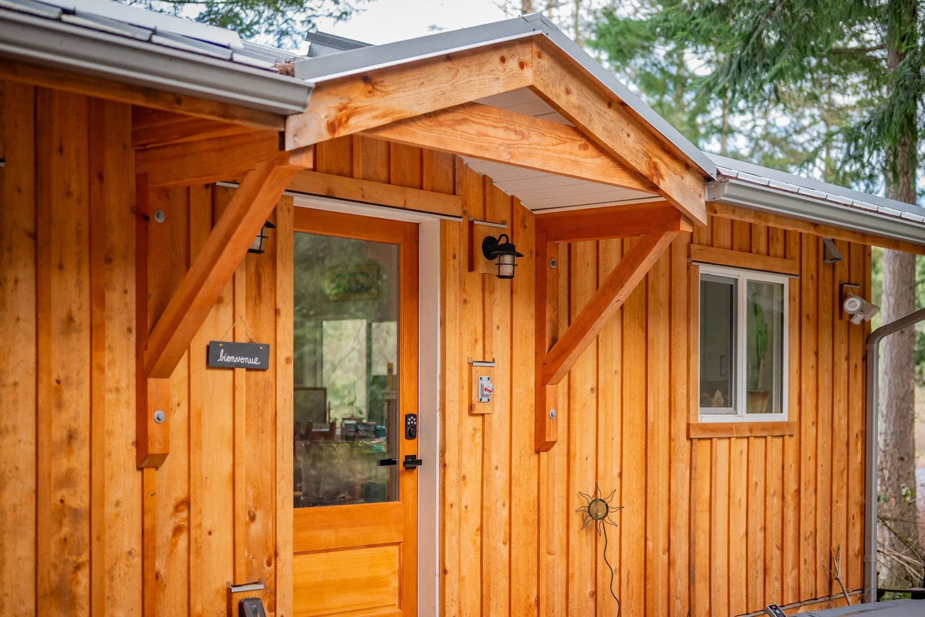 Wooden house exterior with a glass front door, a small window, and outdoor lights. Signs and decorative items are attached to the wooden siding.