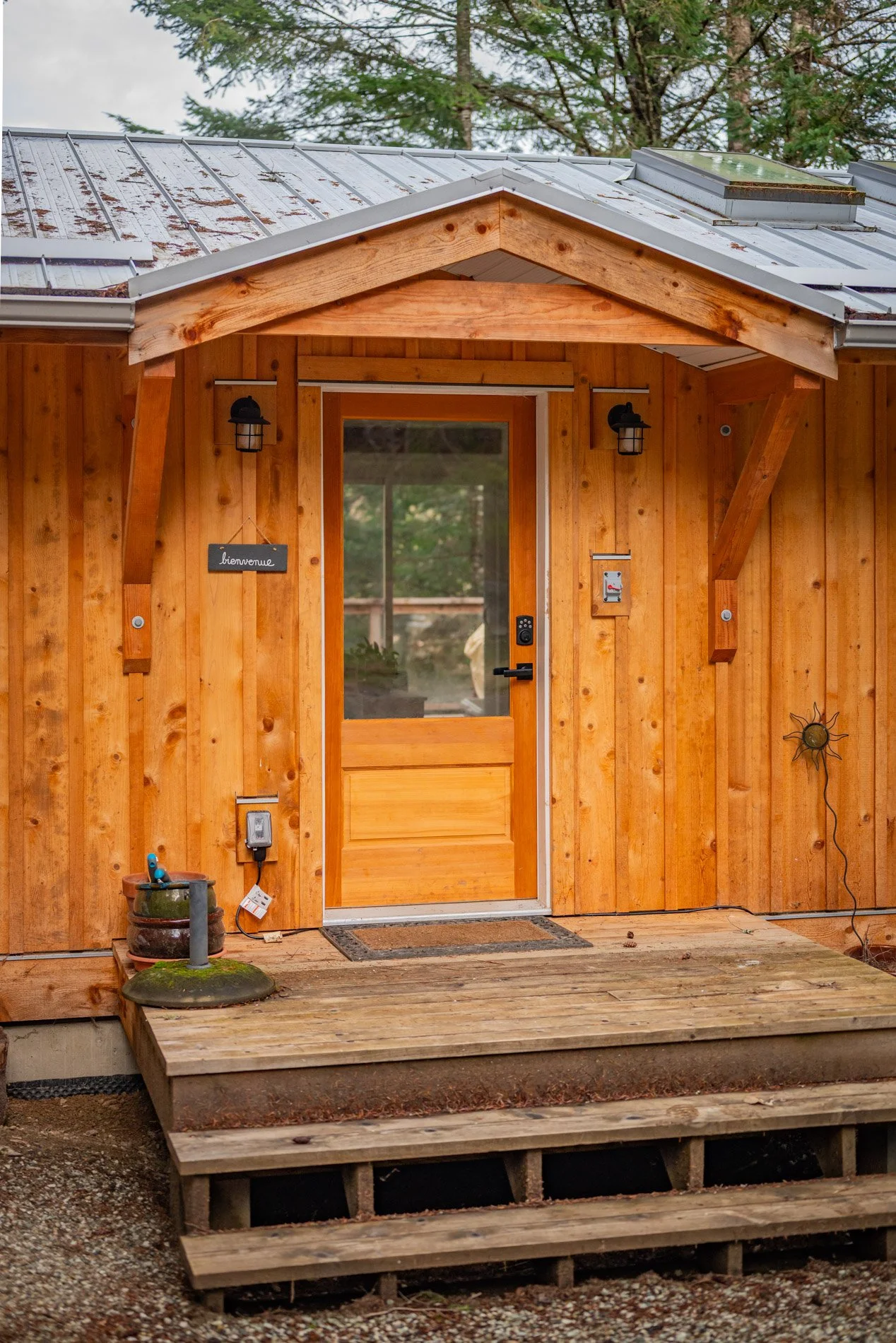 Front view of a small wooden house with a metal roof, steps leading up to the door, and various outdoor decorations and lighting.