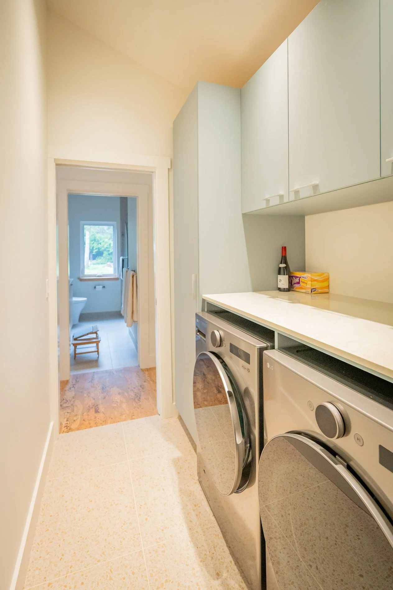 Laundry room with washer and dryer, upper cabinets, and a countertop, with an open doorway leading to a bathroom with a window and towels.