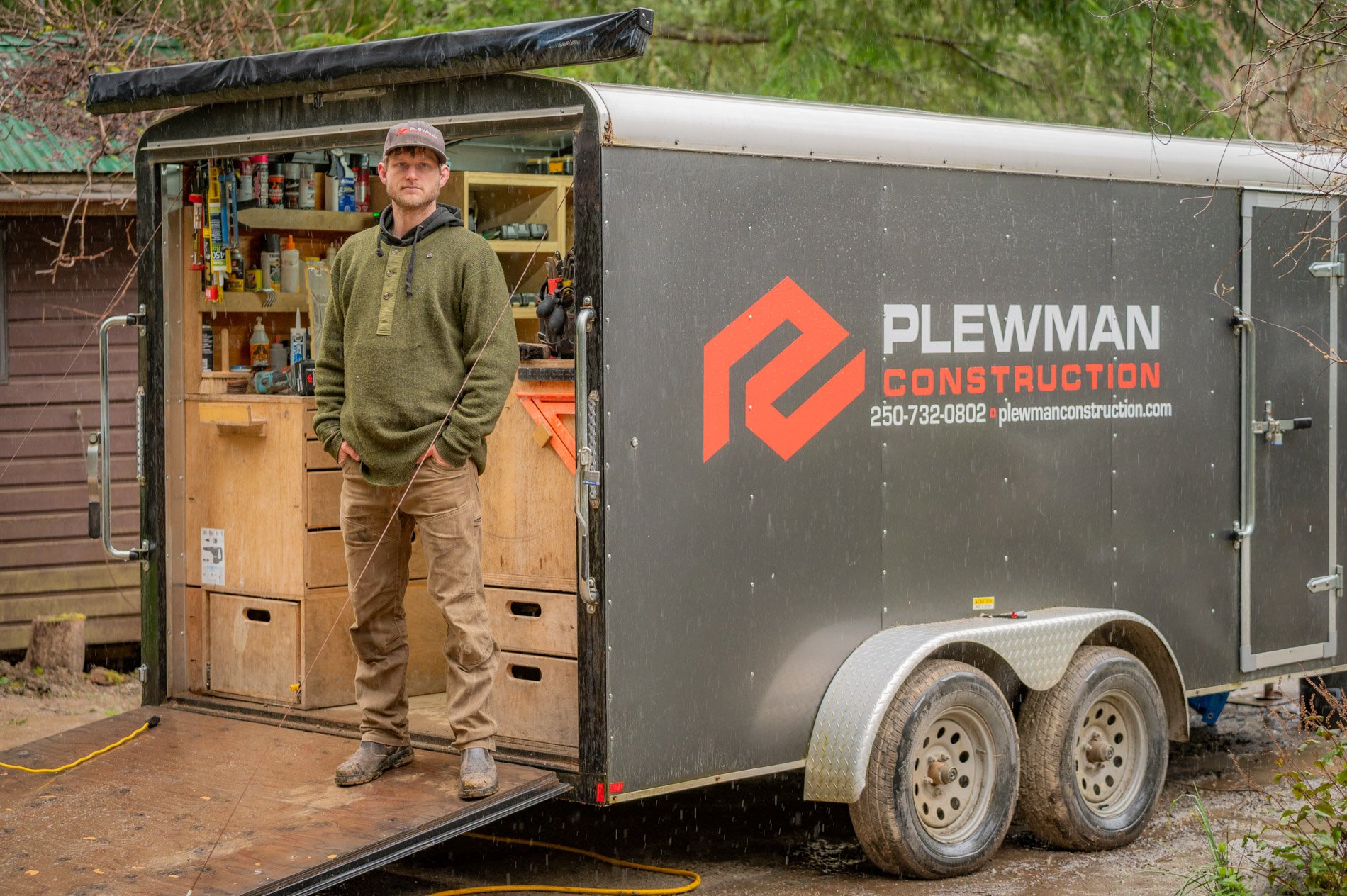 A man standing inside a mobile woodwork or construction workshop trailer, with shelves of tools, and a person in work clothes, parked outdoors on a muddy ground surrounded by trees and wooden structures.