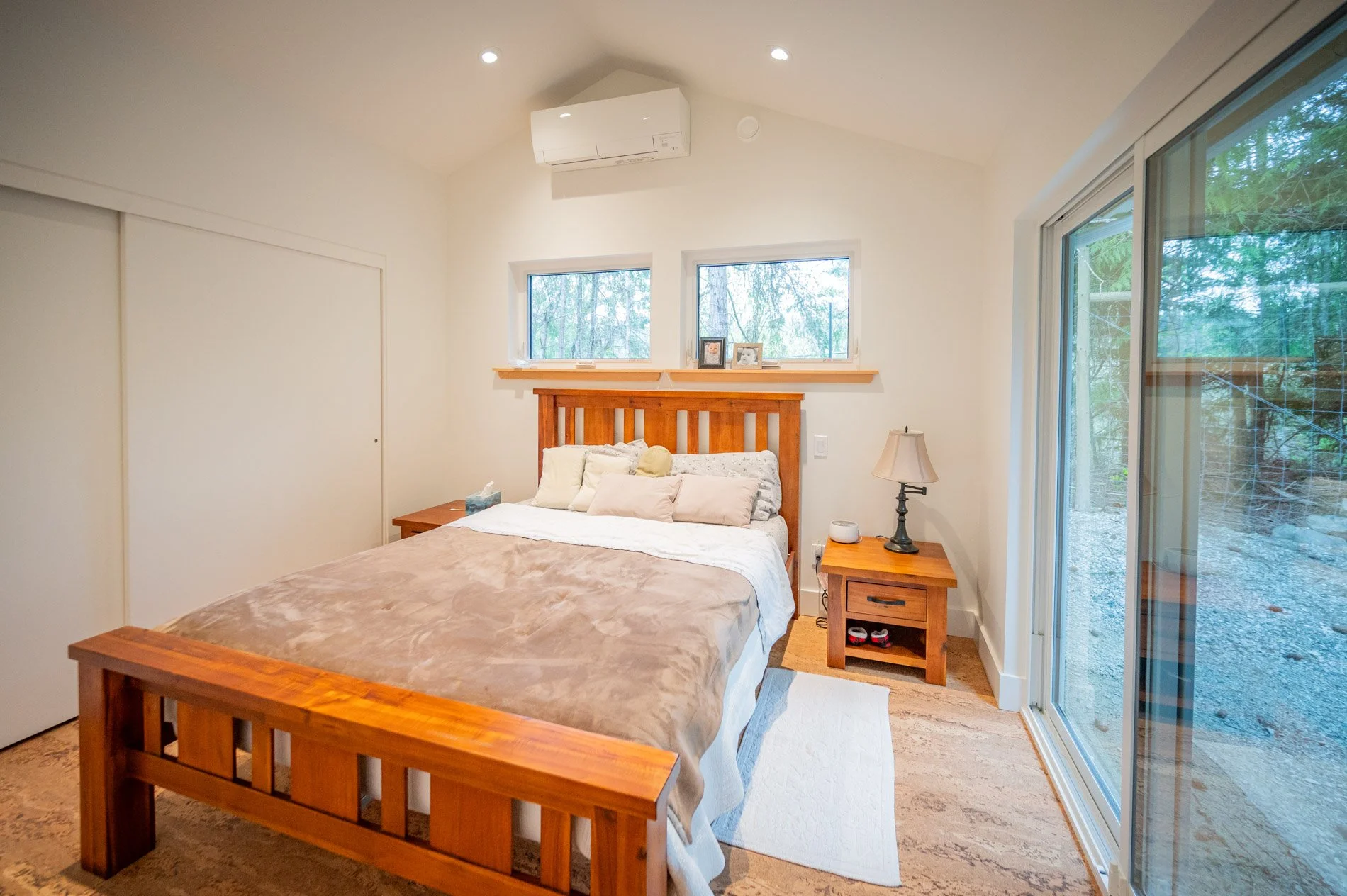 A bedroom with a wooden bed frame, beige blanket, multiple pillows, two windows behind the headboard, a nightstand with a lamp, a white rug, a large sliding glass door, and an air conditioner on the wall.