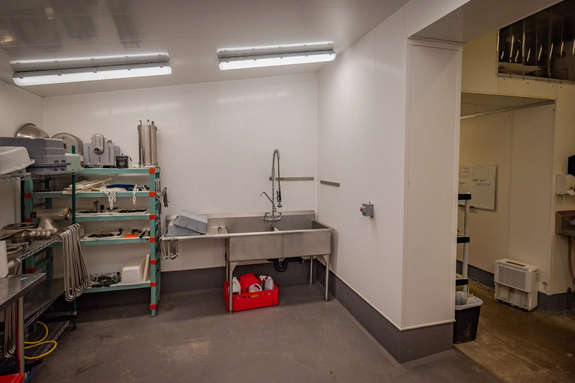 Industrial cleaning or utility room with stainless steel sink, white walls, shelves with various tools and equipment, and fluorescent ceiling lights.