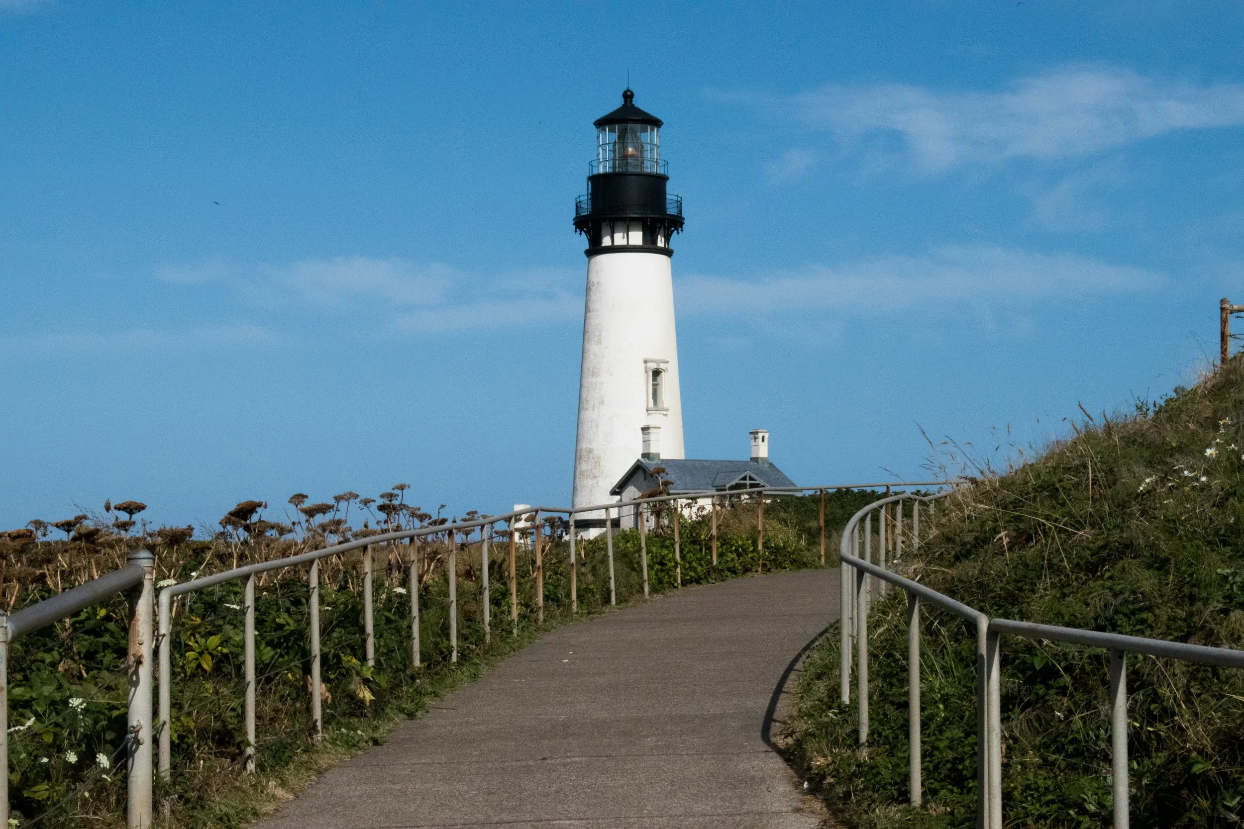 A lighthouse standing on a grassy hill next to a paved walking path with metal railings, against a bright blue sky.