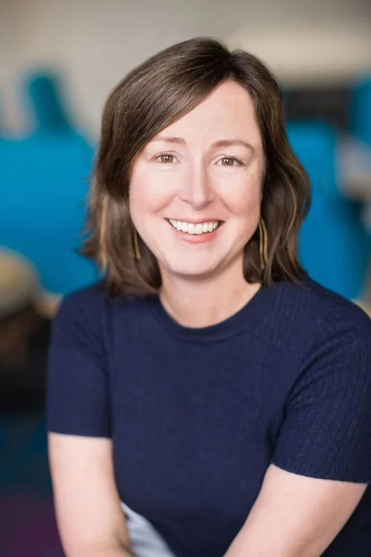 A woman with shoulder-length brown hair smiling, wearing a dark blue top, in an indoor setting with a blurred background.