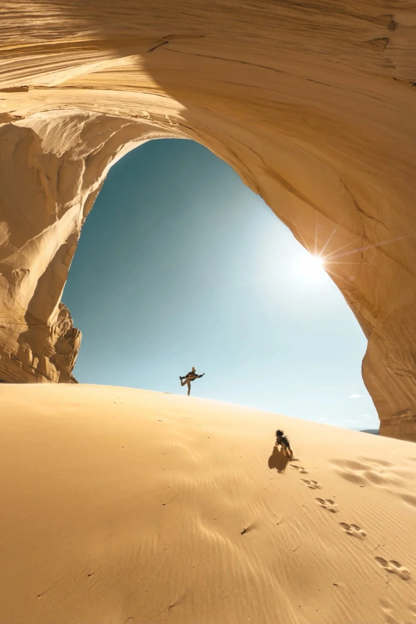A person jumping in a desert landscape with large rock formations and sand dunes, with a bright sun in the sky.