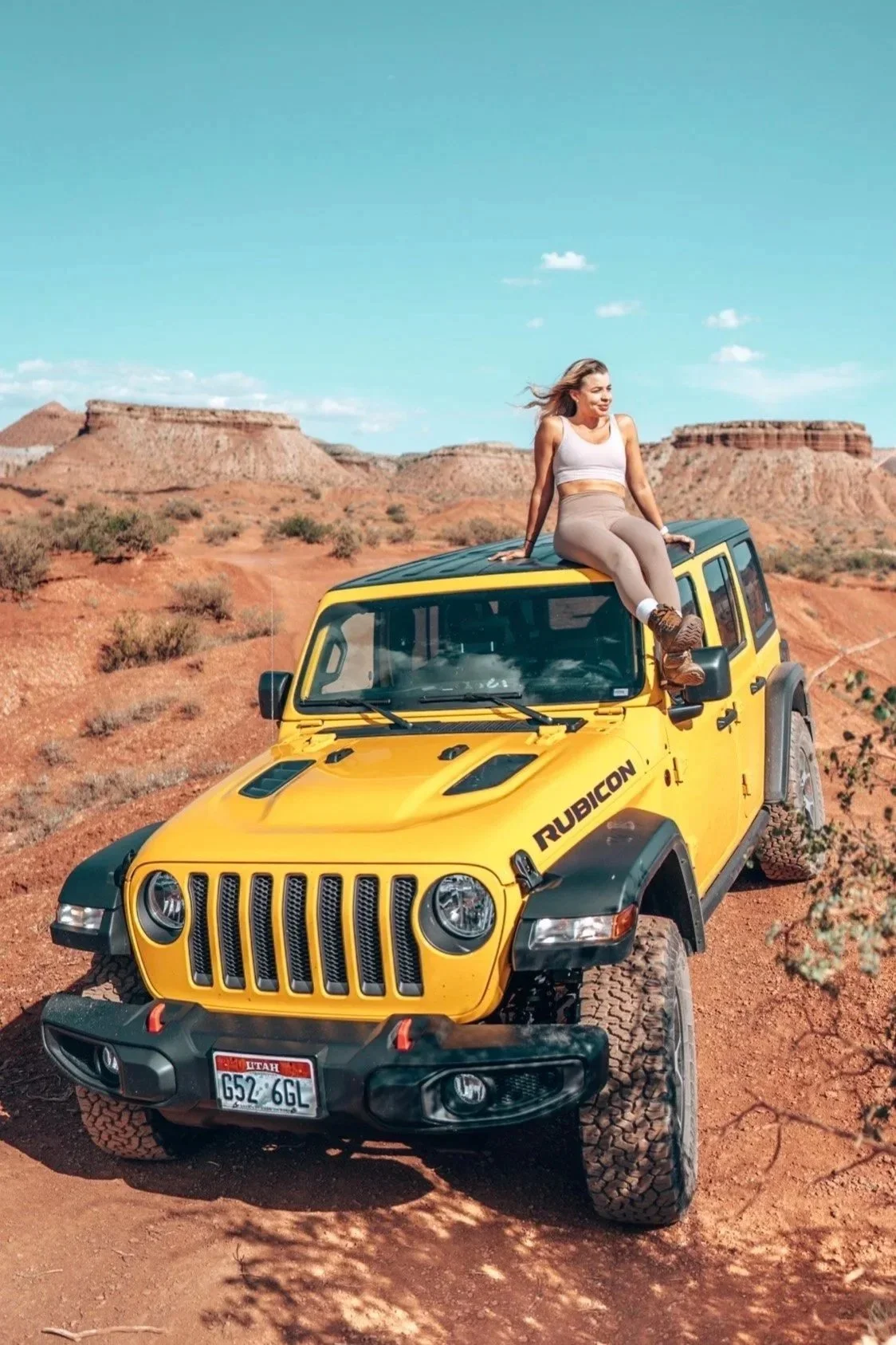 A woman sitting on the roof of a yellow Jeep Rubicon in a desert landscape with rock formations and a blue sky.