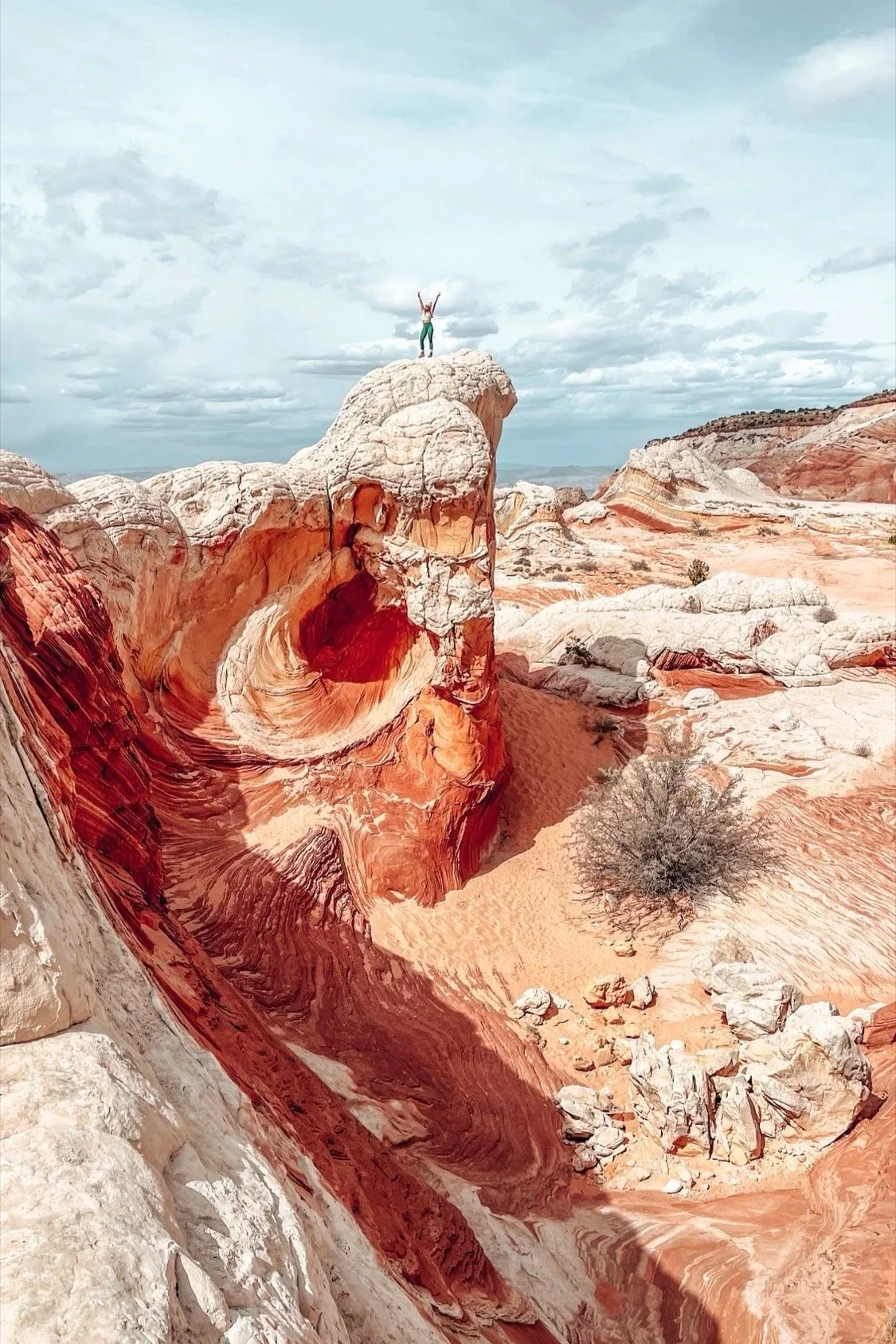 A person standing on top of a large colorful rock formation in a desert landscape with red, white, and orange hues, under a partly cloudy sky.
