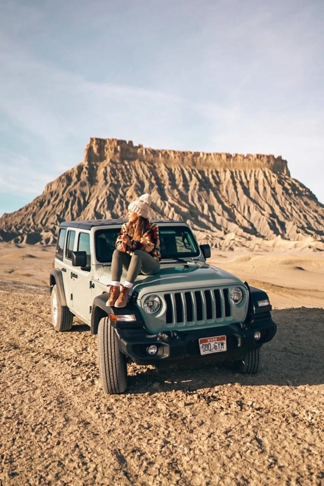 A woman sitting on the hood of a Jeep in a desert with a large flat-topped mountain in the background during sunset.
