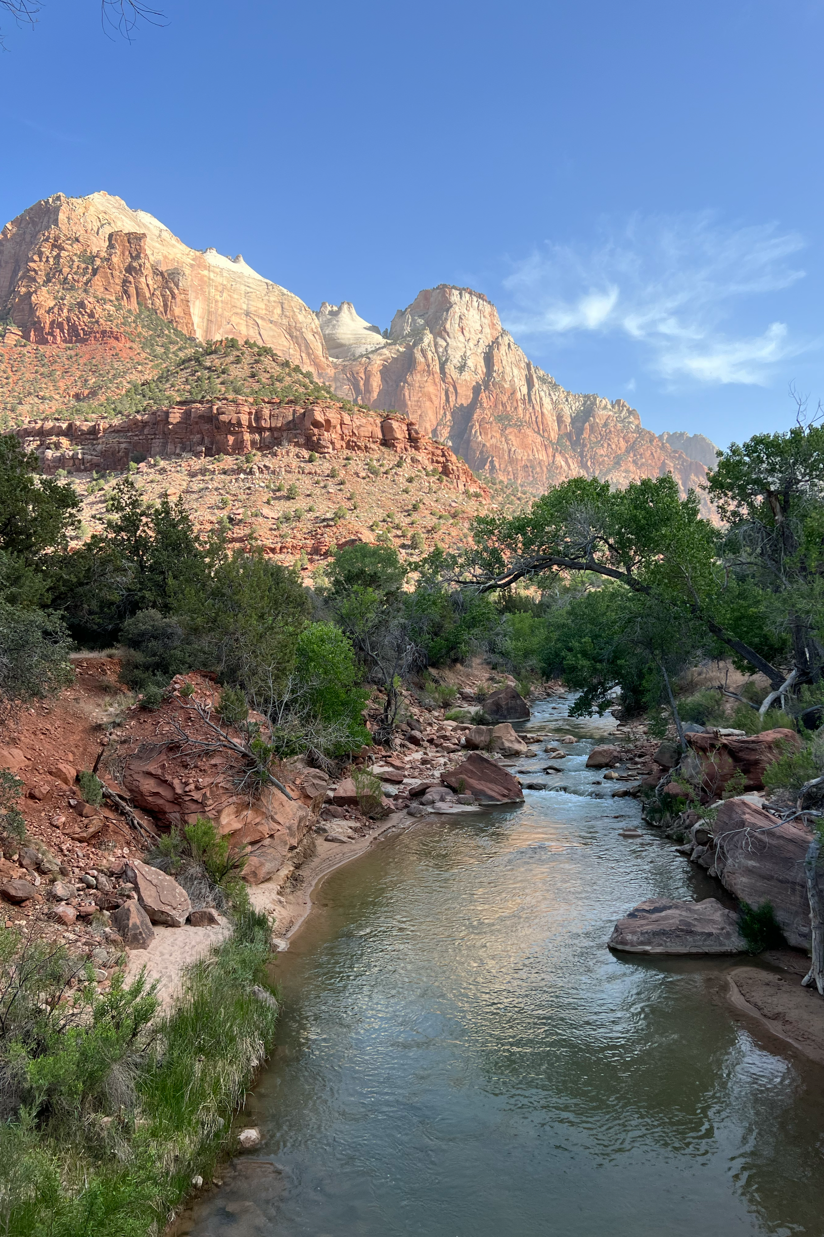 A mountainous landscape with red and orange rock formations, green vegetation, and a small river flowing through the valley, under a clear blue sky with some wispy clouds.